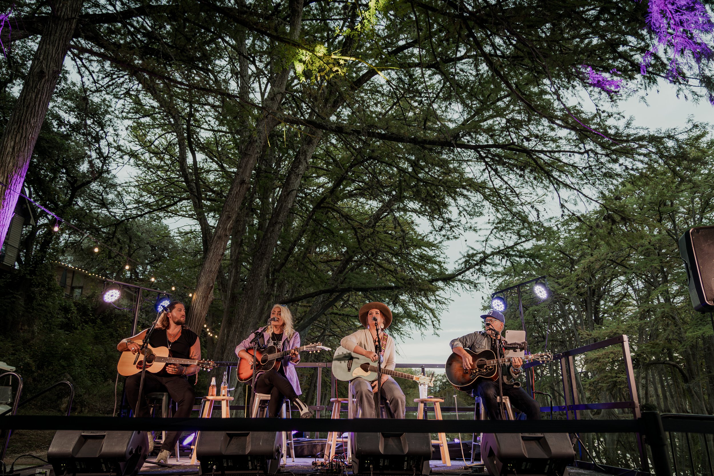 Four musicians perform on an outdoor stage surrounded by trees, each playing a guitar and singing into microphones, with stage lights and speakers visible.