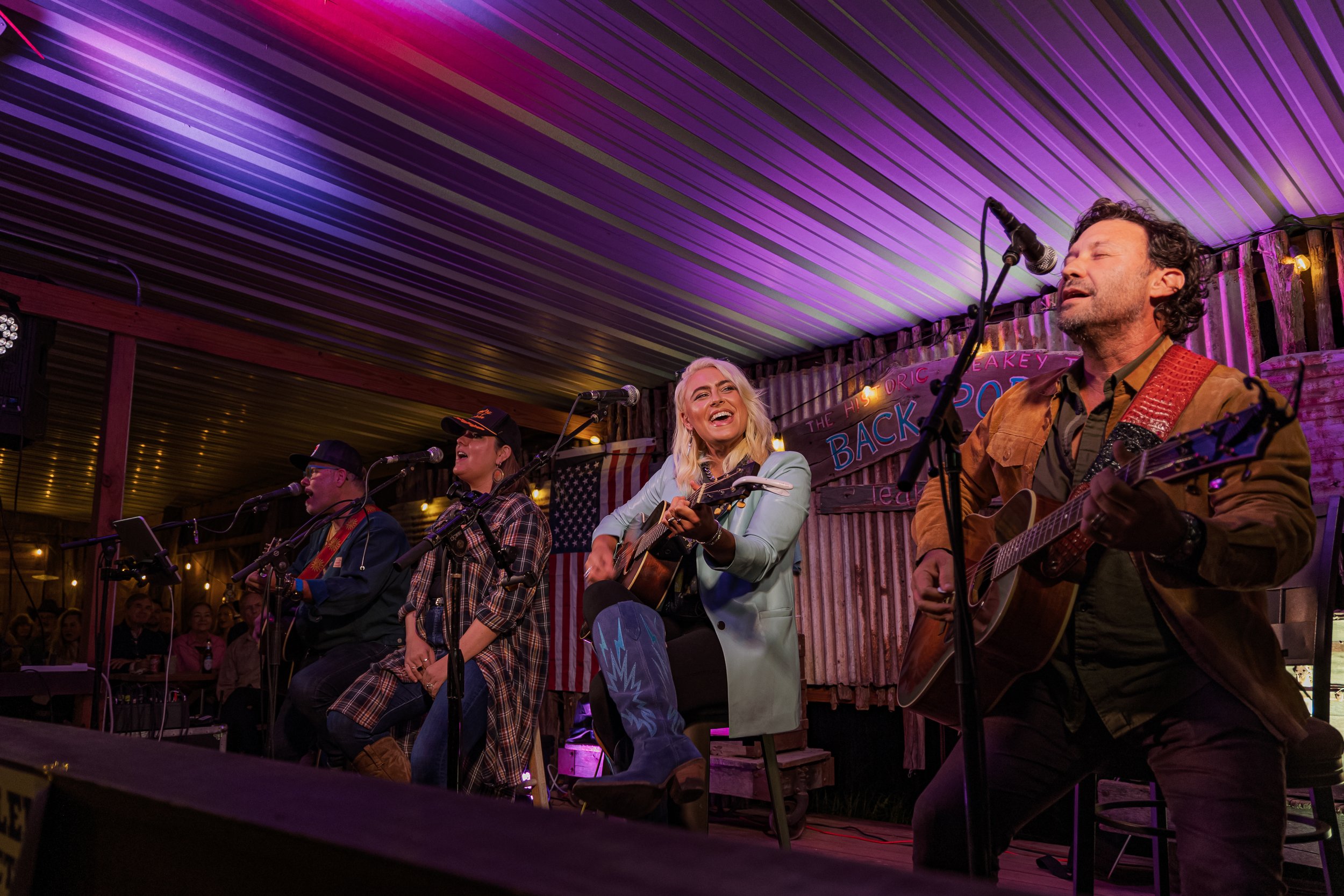 A team of four musicians perform on stage at a country music event. The woman in the center is singing and playing an acoustic guitar, smiling and wearing a light blue coat. The man on the right is playing an acoustic guitar and singing into a microp