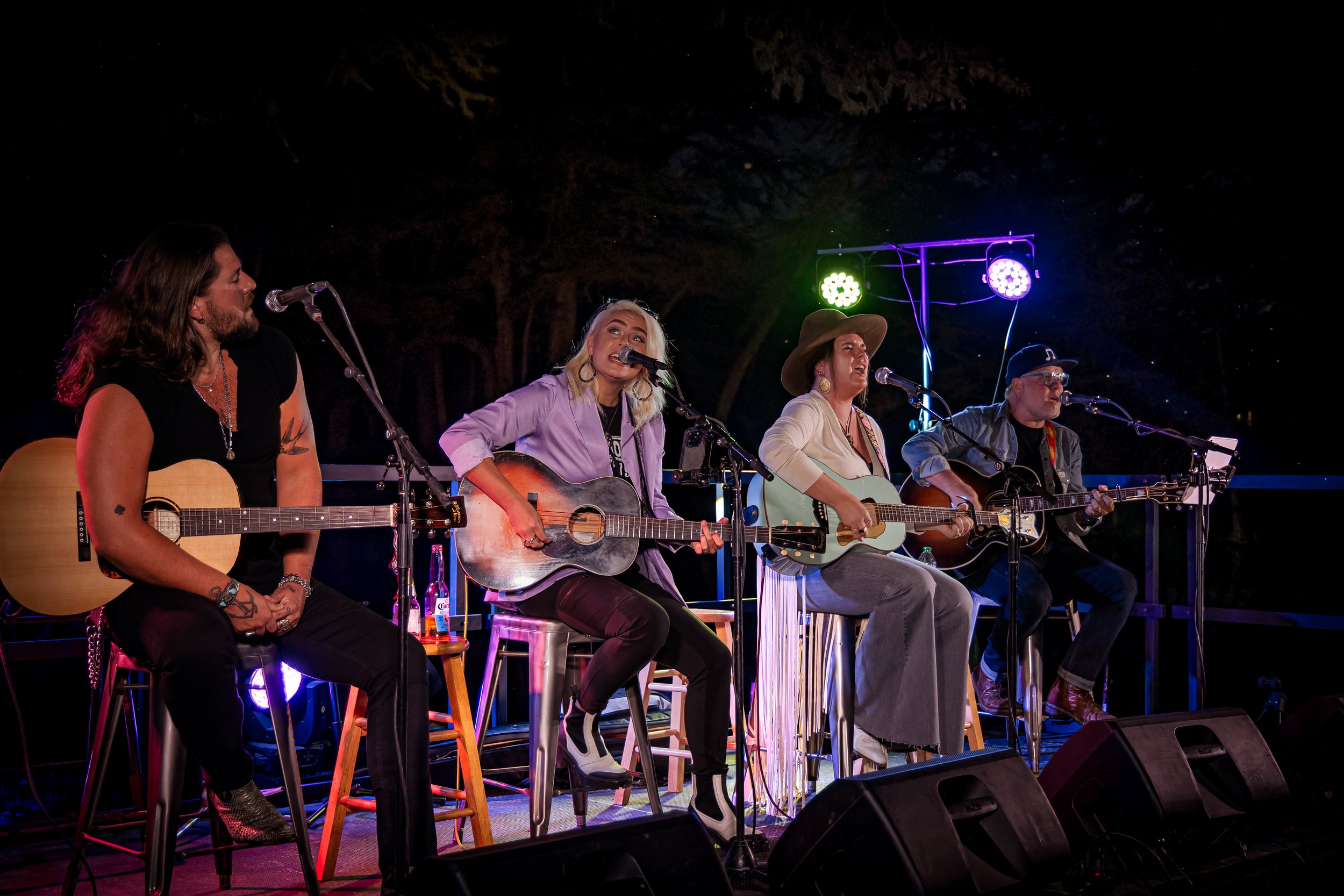 Four musicians playing guitars on stage during a night performance, with colorful stage lighting and trees in the background.