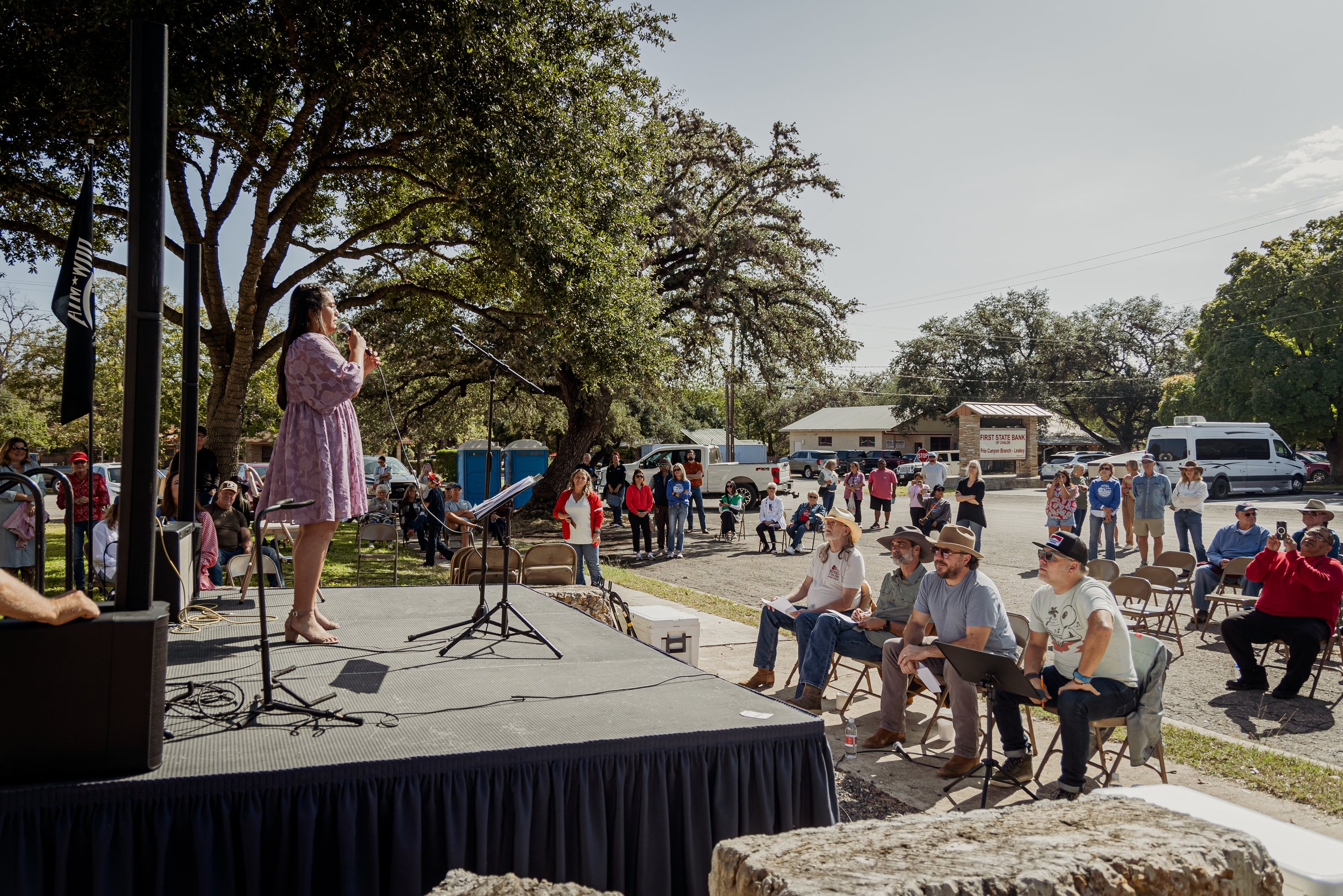 A woman in a pink dress speaking into a microphone on an outdoor stage during a community event, with an audience seated and standing nearby on a sunny day