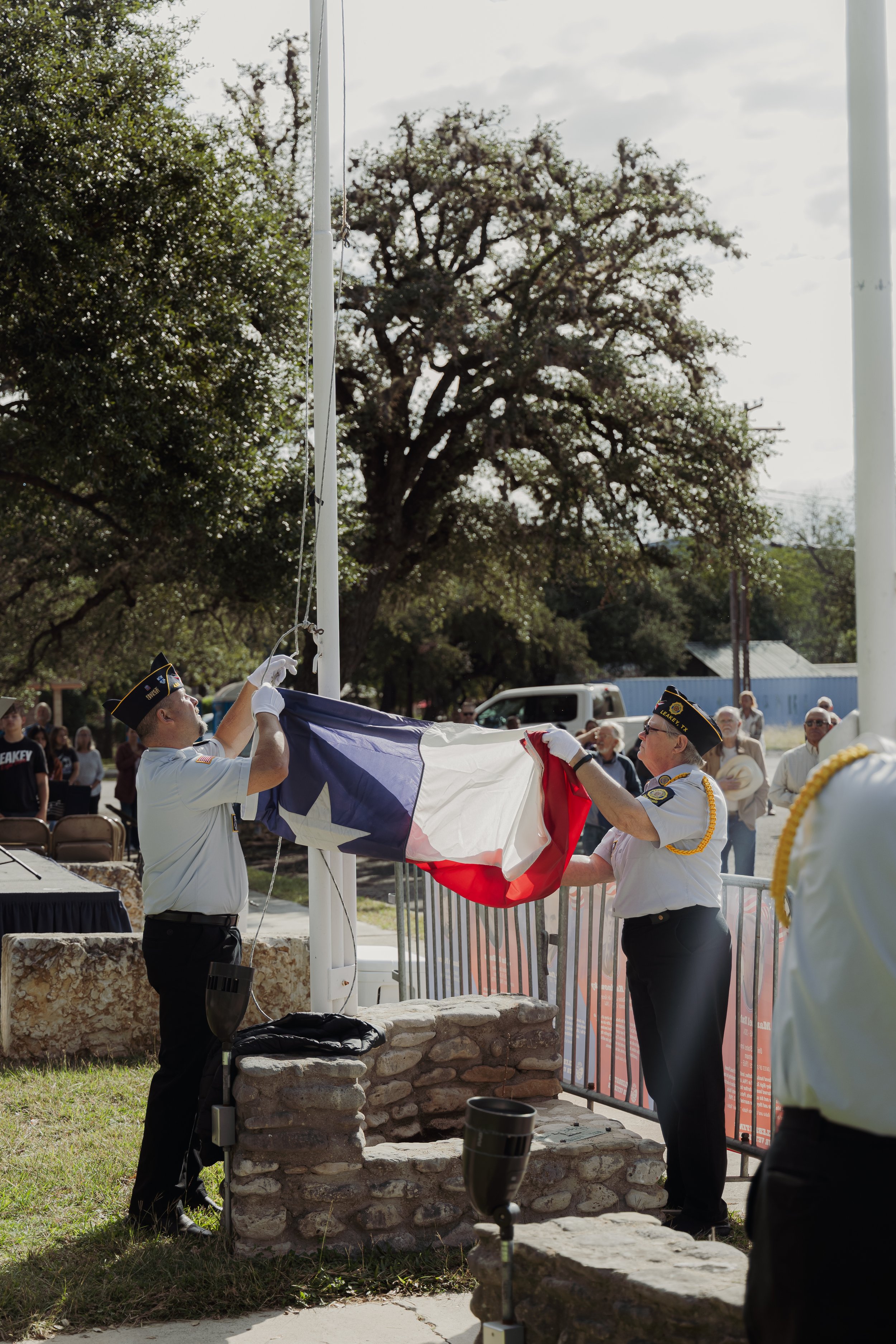 Two uniformed veterans are folding the Texas state flag during a public ceremony, with spectators watching in the background.