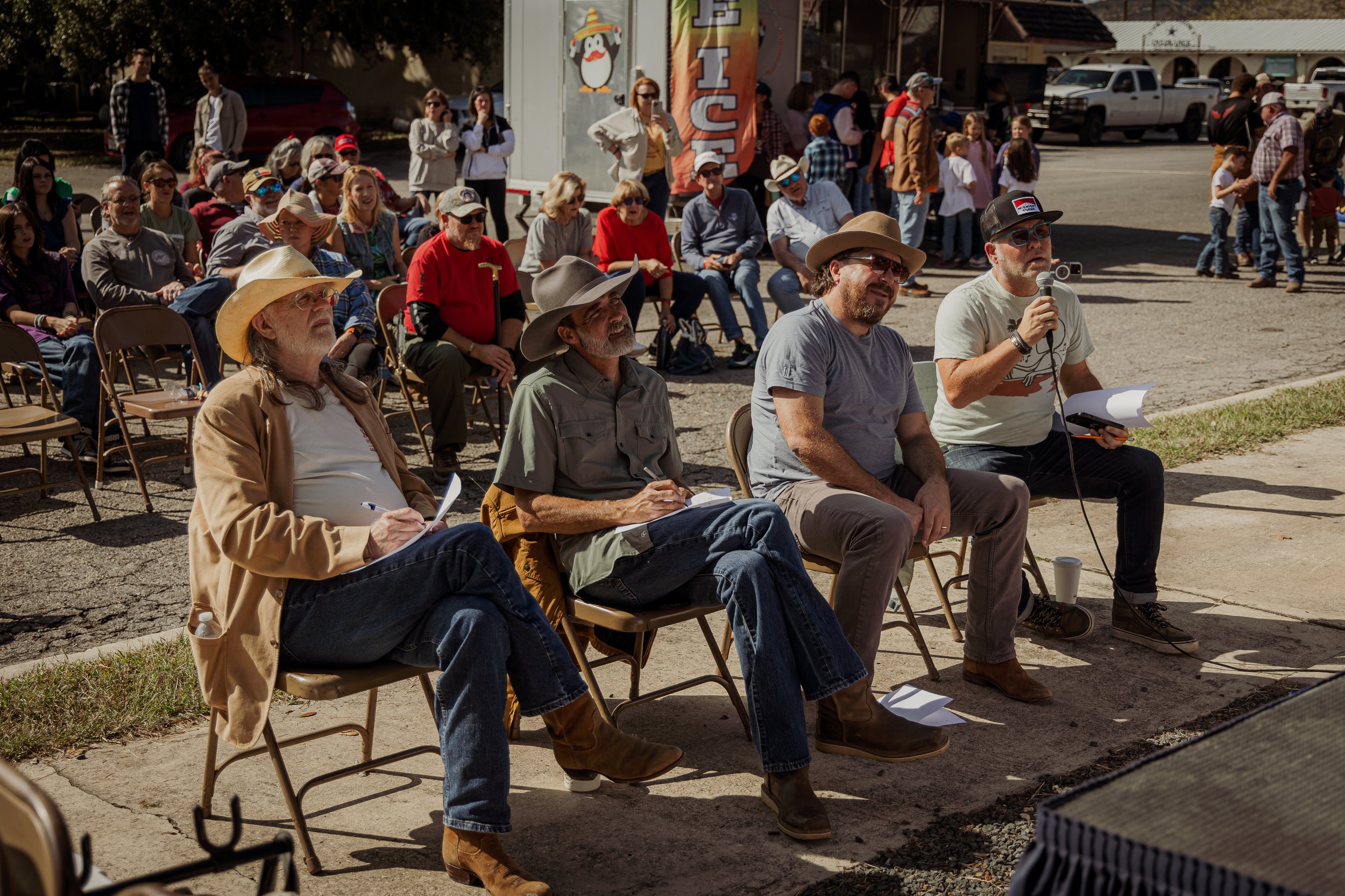Four men sitting outdoors during a daytime event, with one speaking into a microphone, and a crowd of people seated and standing behind them.