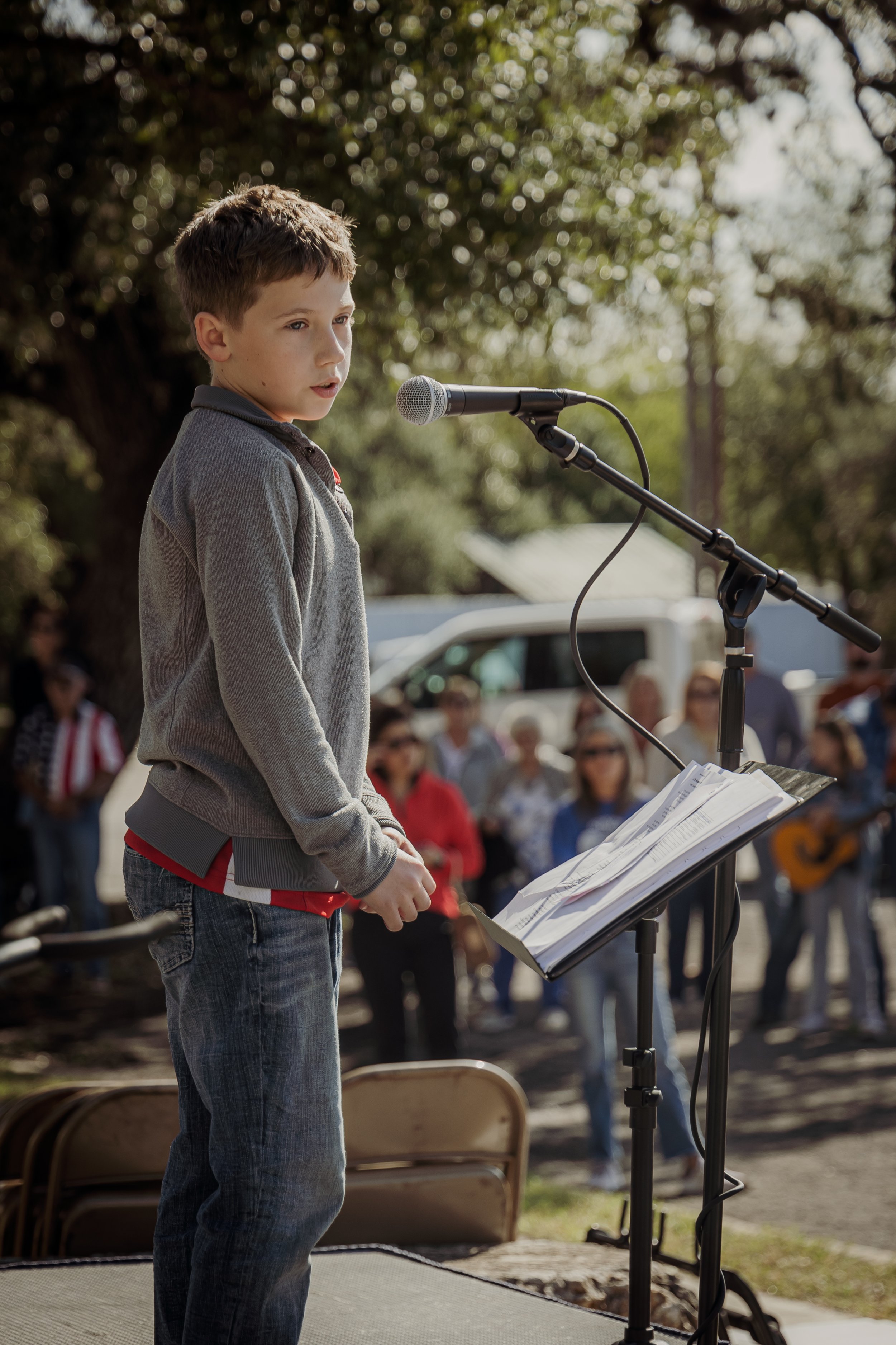 Young boy speaking into microphone during outdoor event with an audience watching.