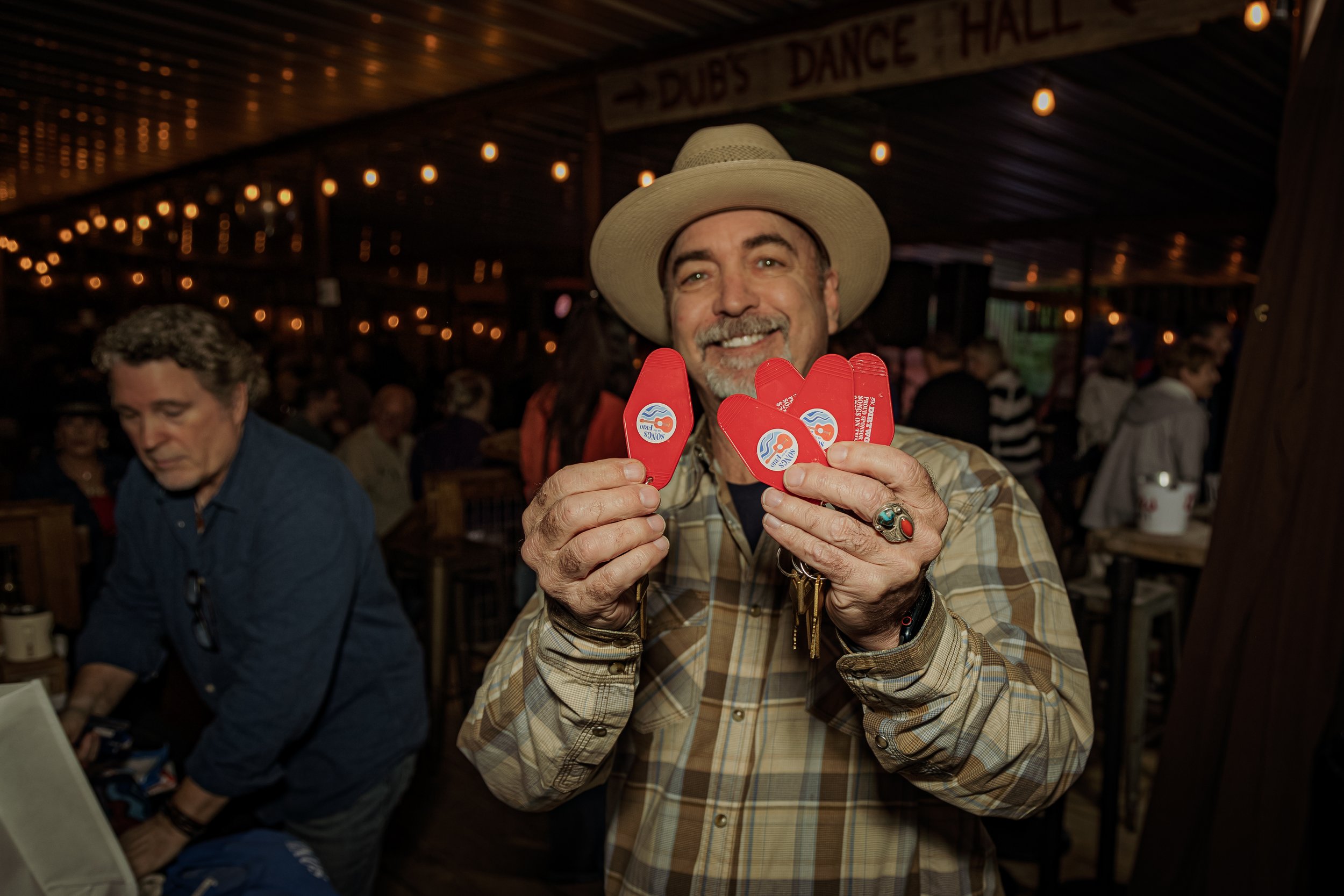 Man smiling and holding several red poker chips inside a lively indoor venue with string lights and people in the background.