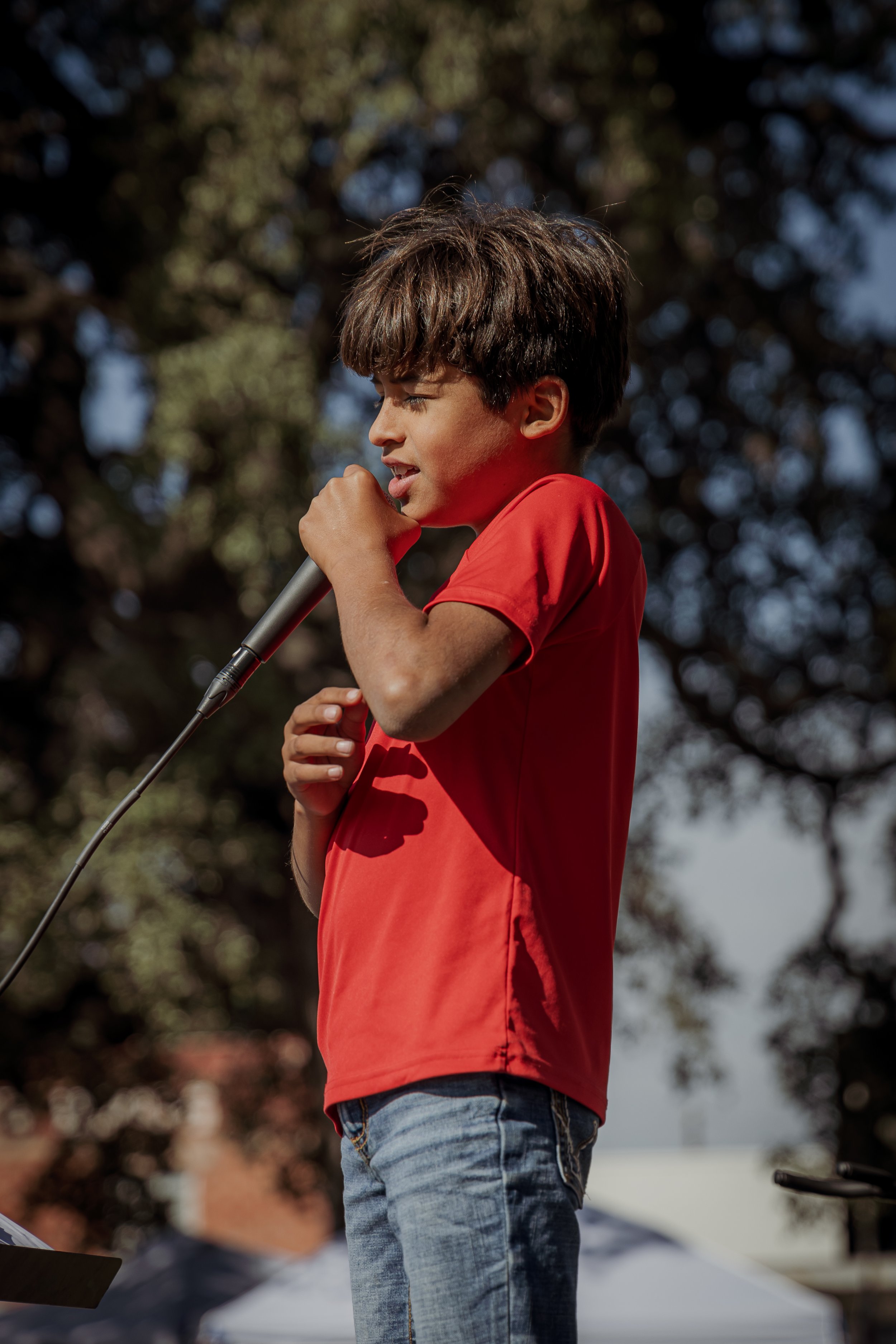 A young boy in a red shirt and jeans holding a microphone, appears to be singing or speaking outdoors during the daytime.