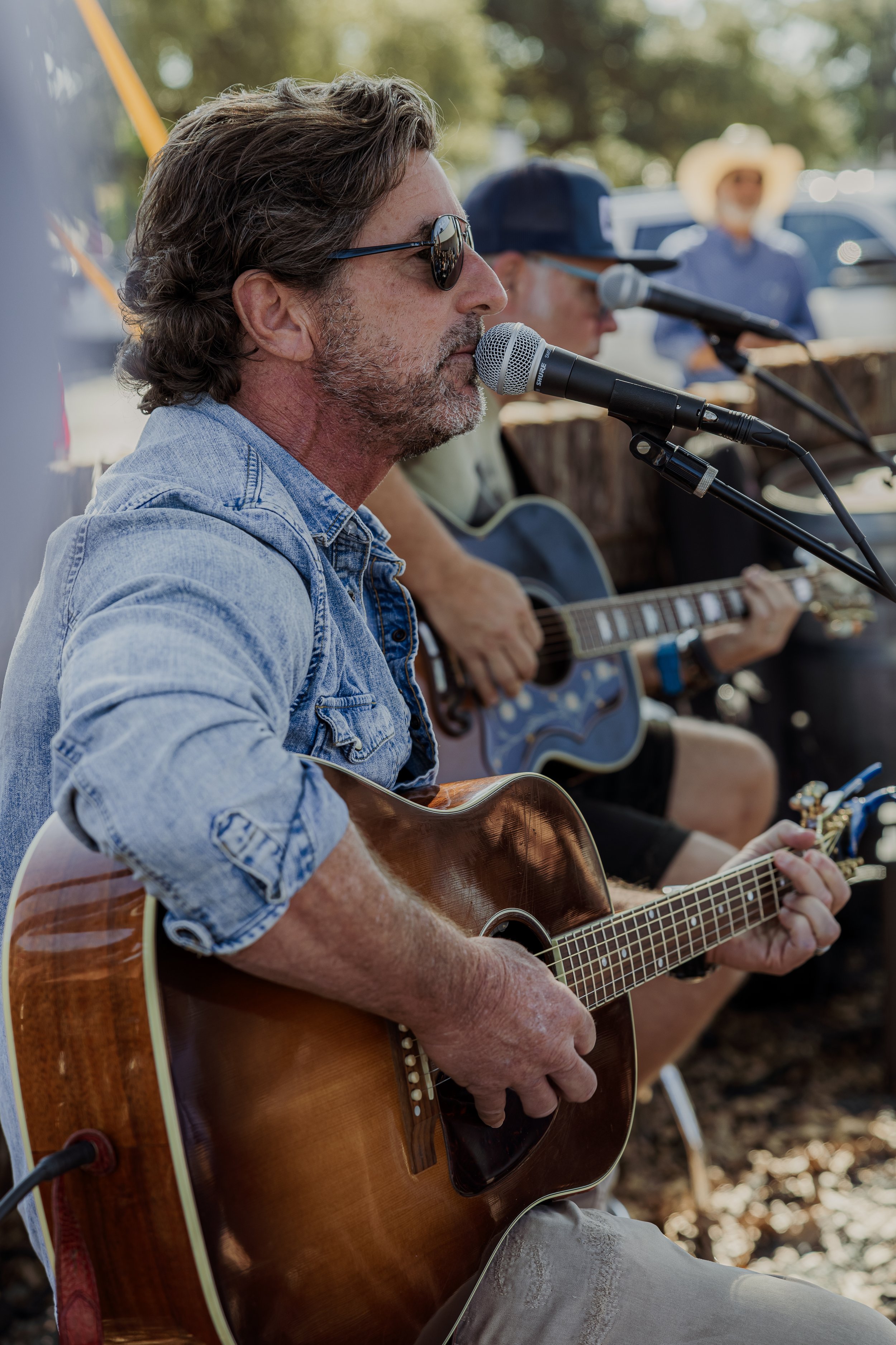 A man with sunglasses and grey hair playing an acoustic guitar and singing into a microphone at an outdoor event, with two other musicians in the background, one playing a guitar and the other wearing a cowboy hat.