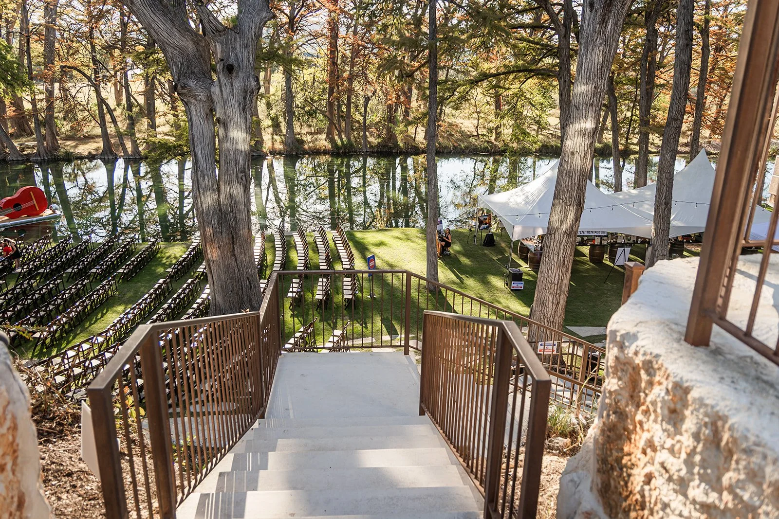 View of outdoor event space with rows of chairs facing a lake, surrounded by trees, with a staircase leading down to the grass and tents on the right
