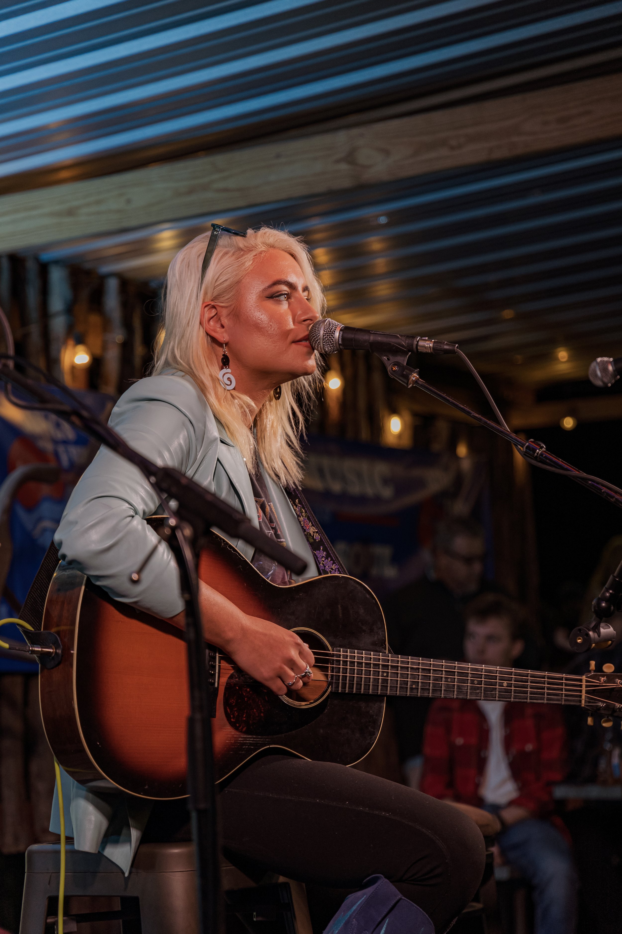 A blonde woman wearing a light blue jacket and earrings is playing an acoustic guitar and singing into a microphone on stage at a music venue.