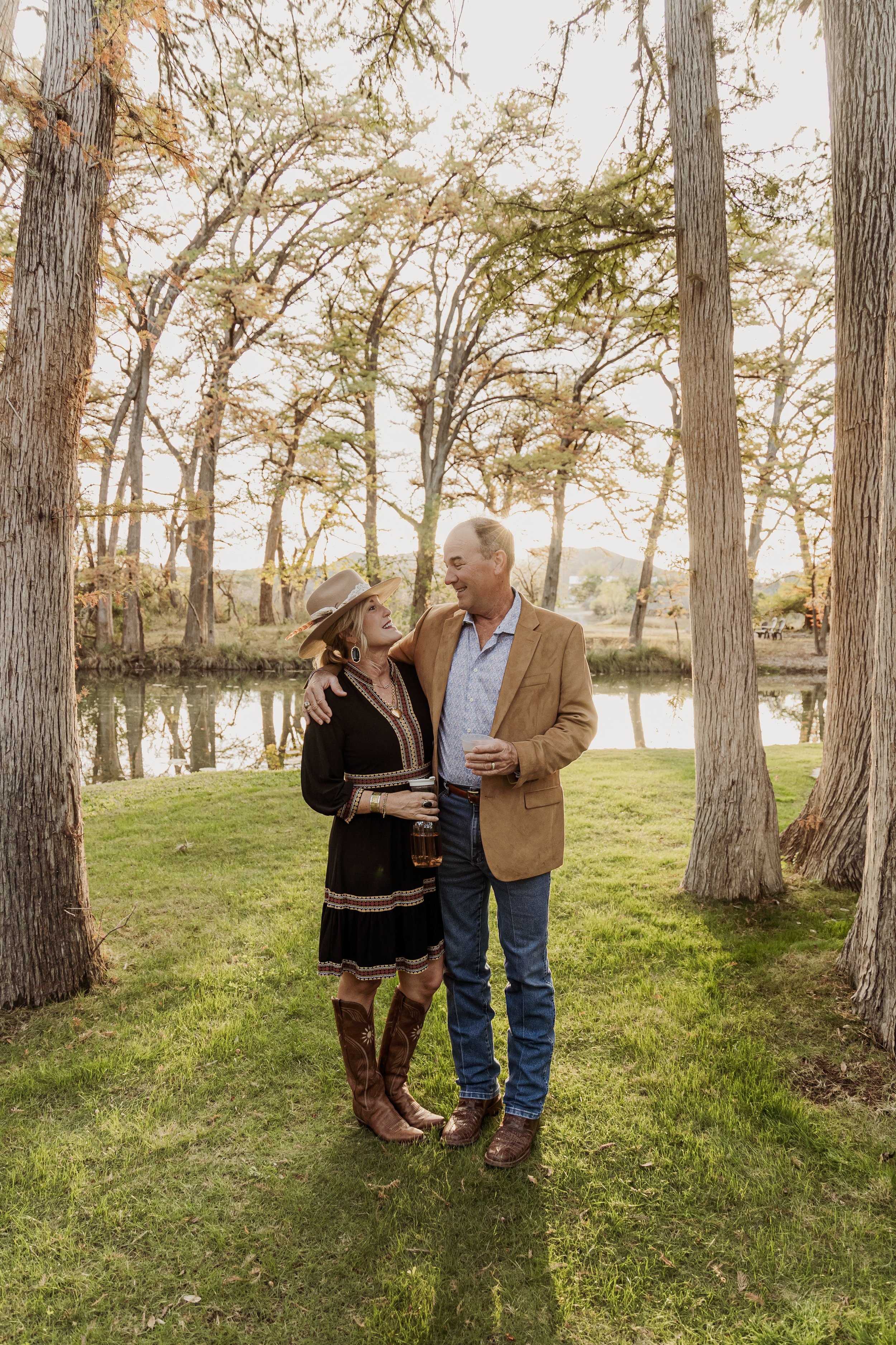 A man and woman standing together outdoors by a pond, smiling and holding drinks, surrounded by trees and grass at sunset.