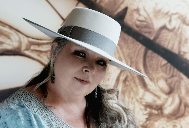 A woman wearing a large white hat with a gray band, gray dress, and earrings.