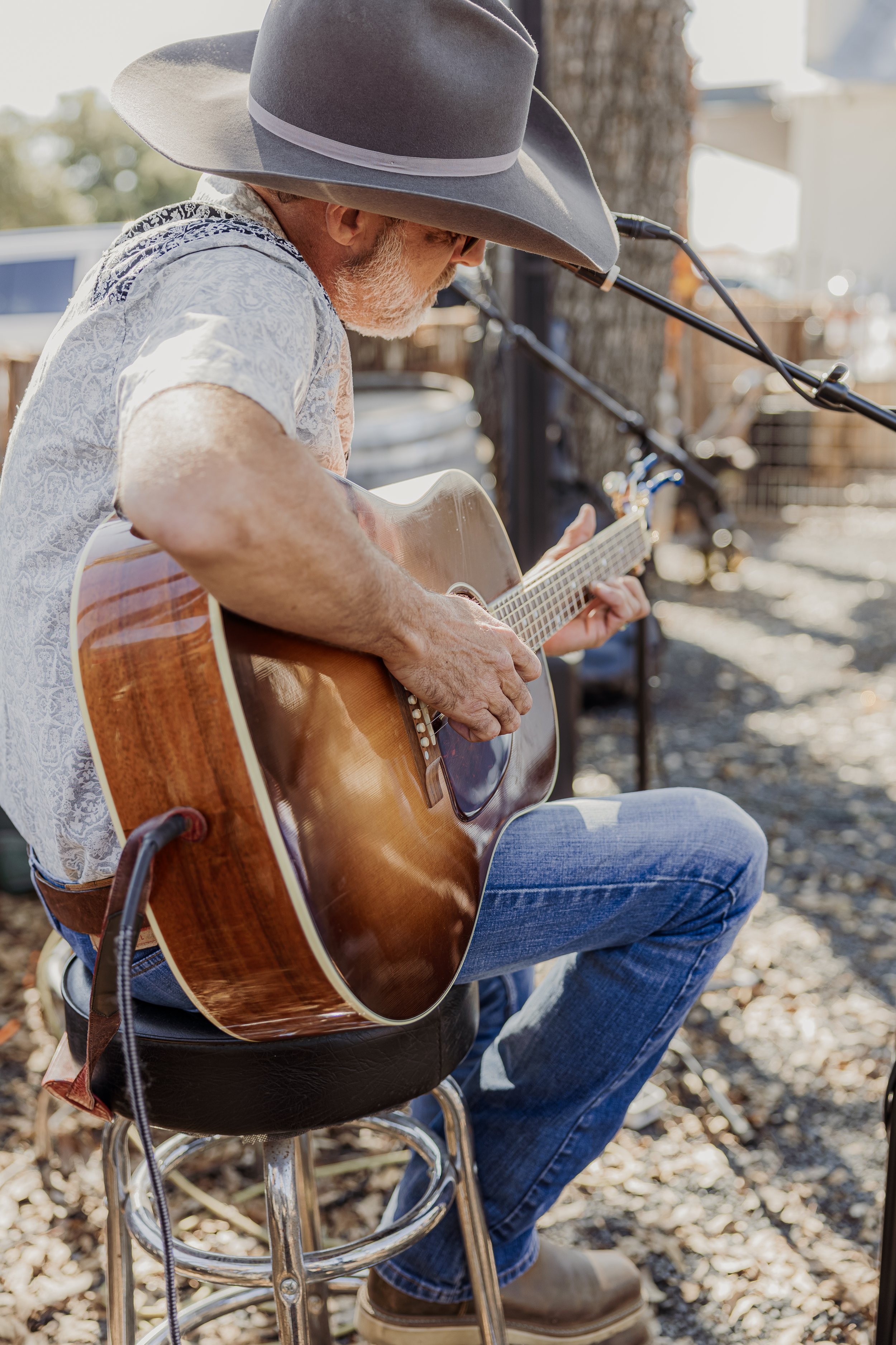 A man wearing a large gray cowboy hat, a patterned short-sleeve shirt, and jeans is sitting on a stool and playing an acoustic guitar outdoors next to a tree, with a microphone setup in front of him.