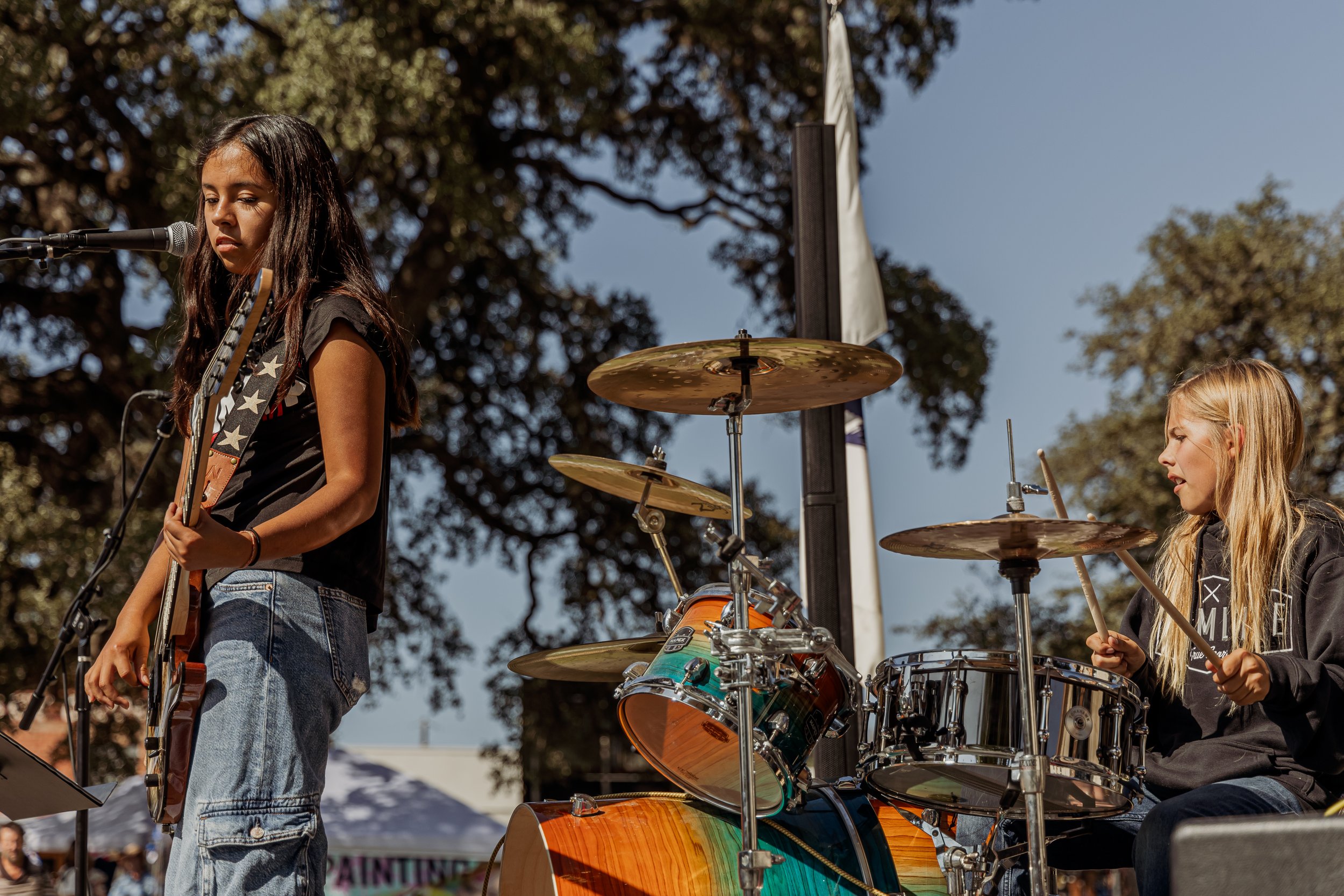 Two young girls performing music outdoors on a sunny day. One girl with long dark hair is singing into a microphone and playing an electric guitar, while the other girl with long blonde hair is playing the drums.