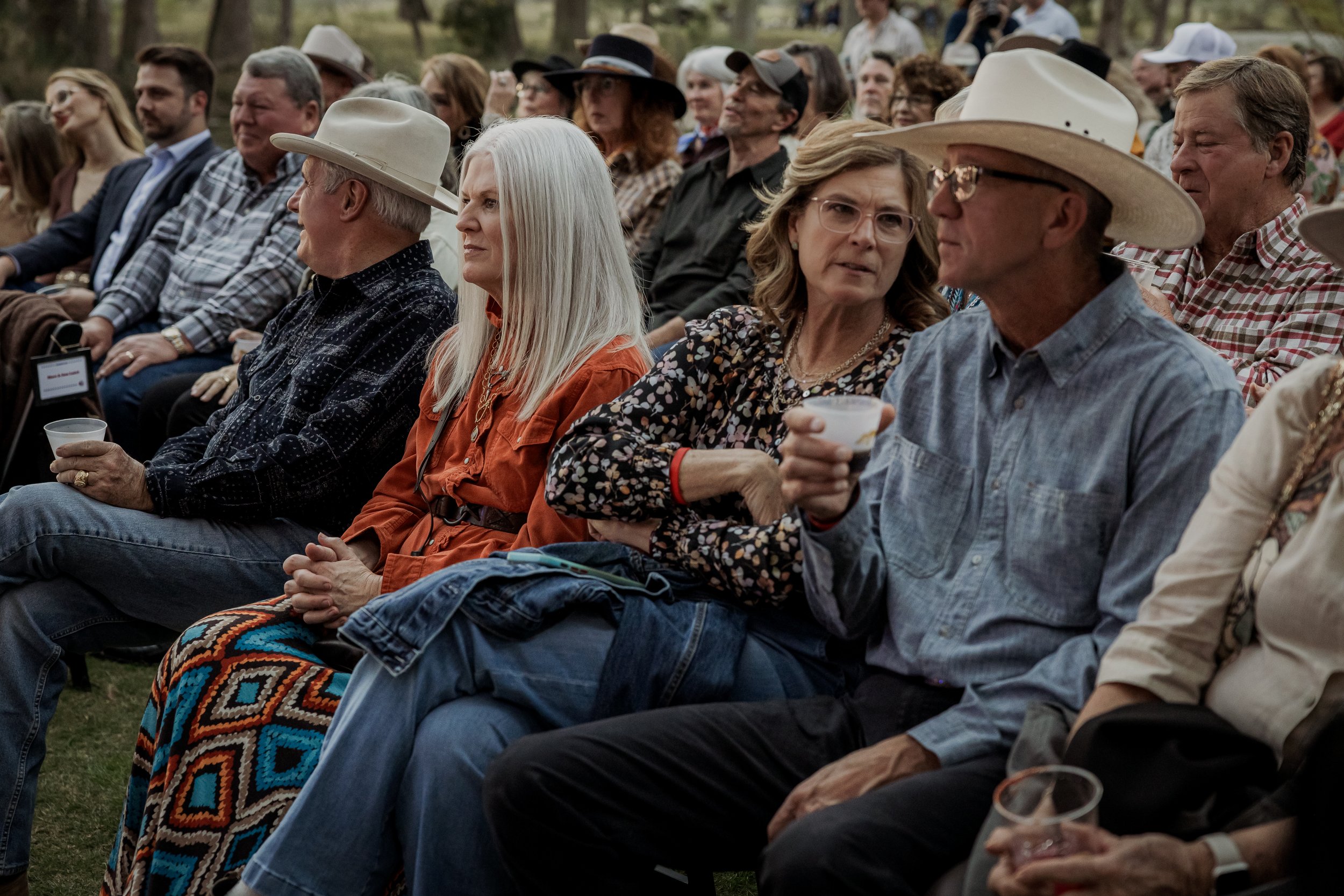 A group of people seated outdoors at an event, some wearing cowboy hats, and some holding cups, engaged in conversation with a crowd in the background.
