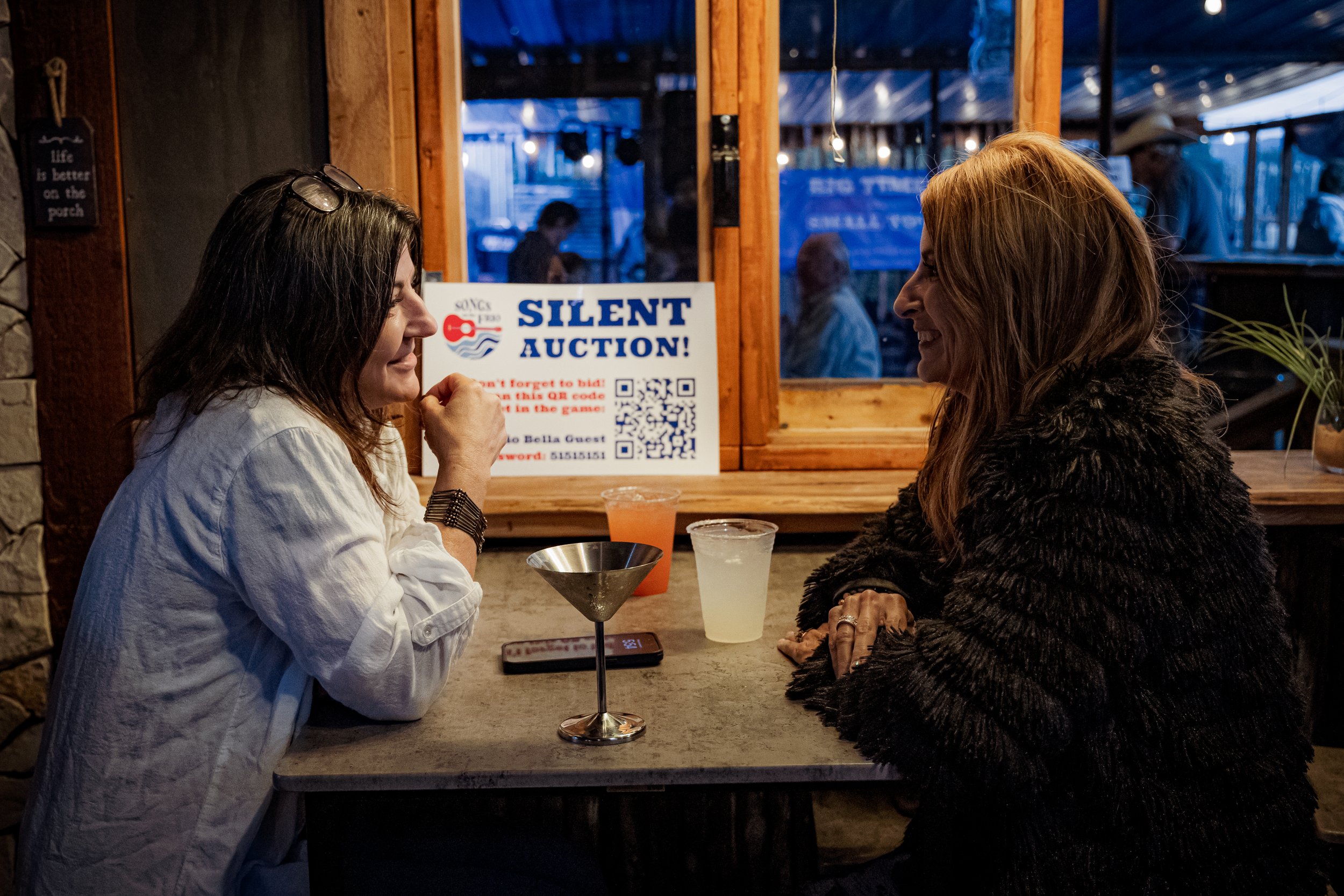 Two women sitting at a bar or restaurant, smiling and engaging in conversation. There are drinks on the table, and a window behind them showing people outside. A sign in the background advertises a silent auction.
