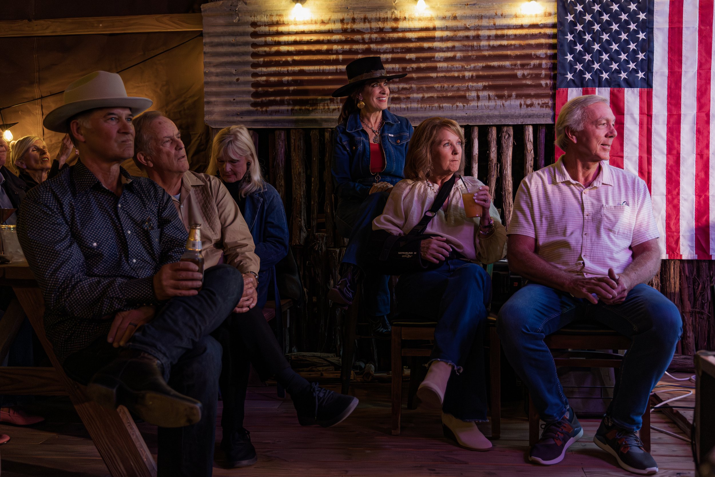 A group of people seated and watching a performance at a rustic venue decorated with American flags, some holding drinks.