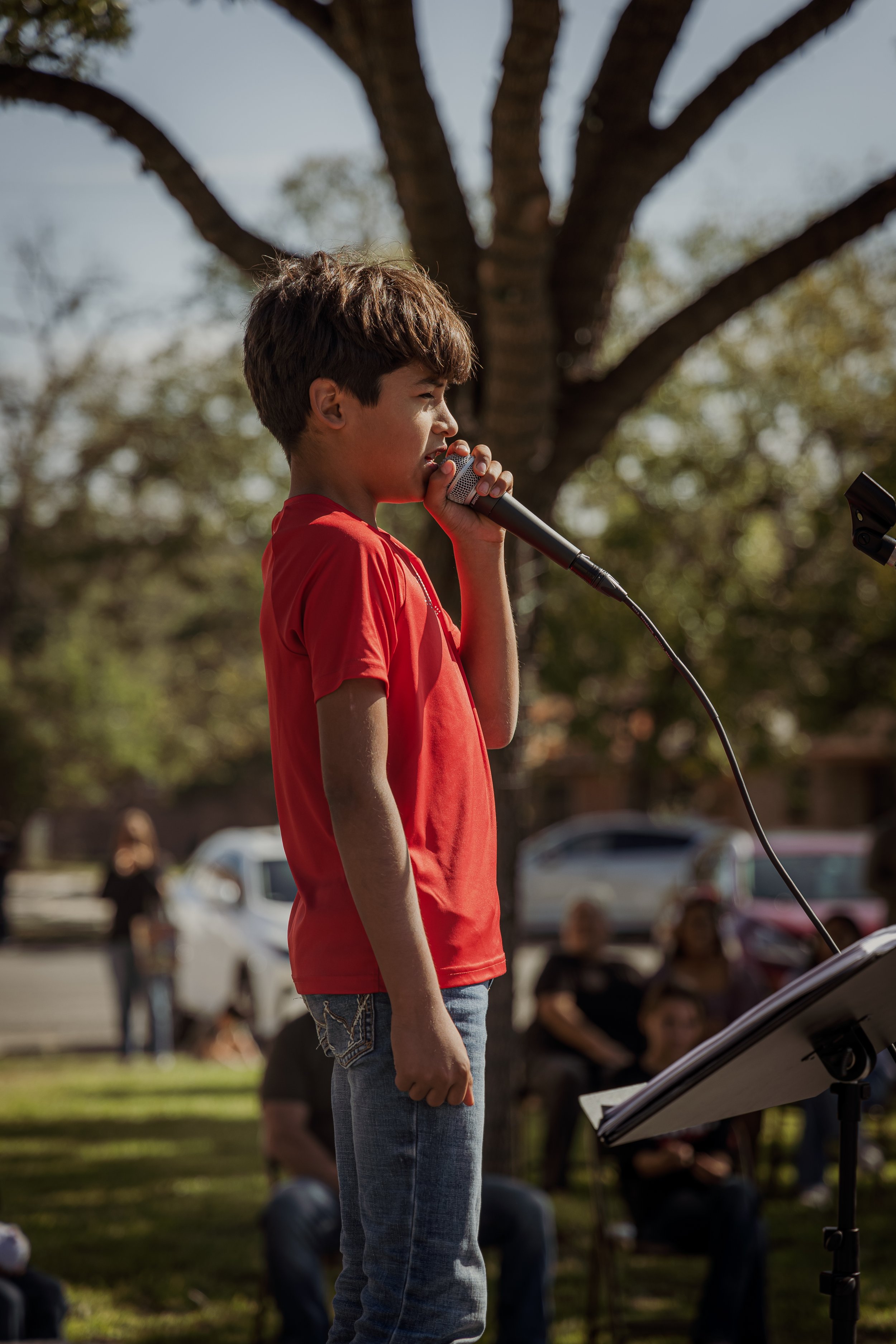 A young boy wearing a red shirt and jeans stands outdoors, singing into a microphone with a stand and music sheet, as people sit and watch in the background during daytime.