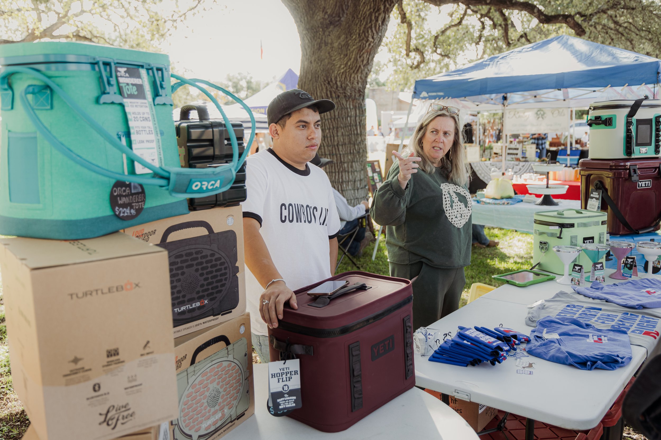 A woman and a man at an outdoor market stall with various branded coolers and merchandise, under a large tree with tents in the background.