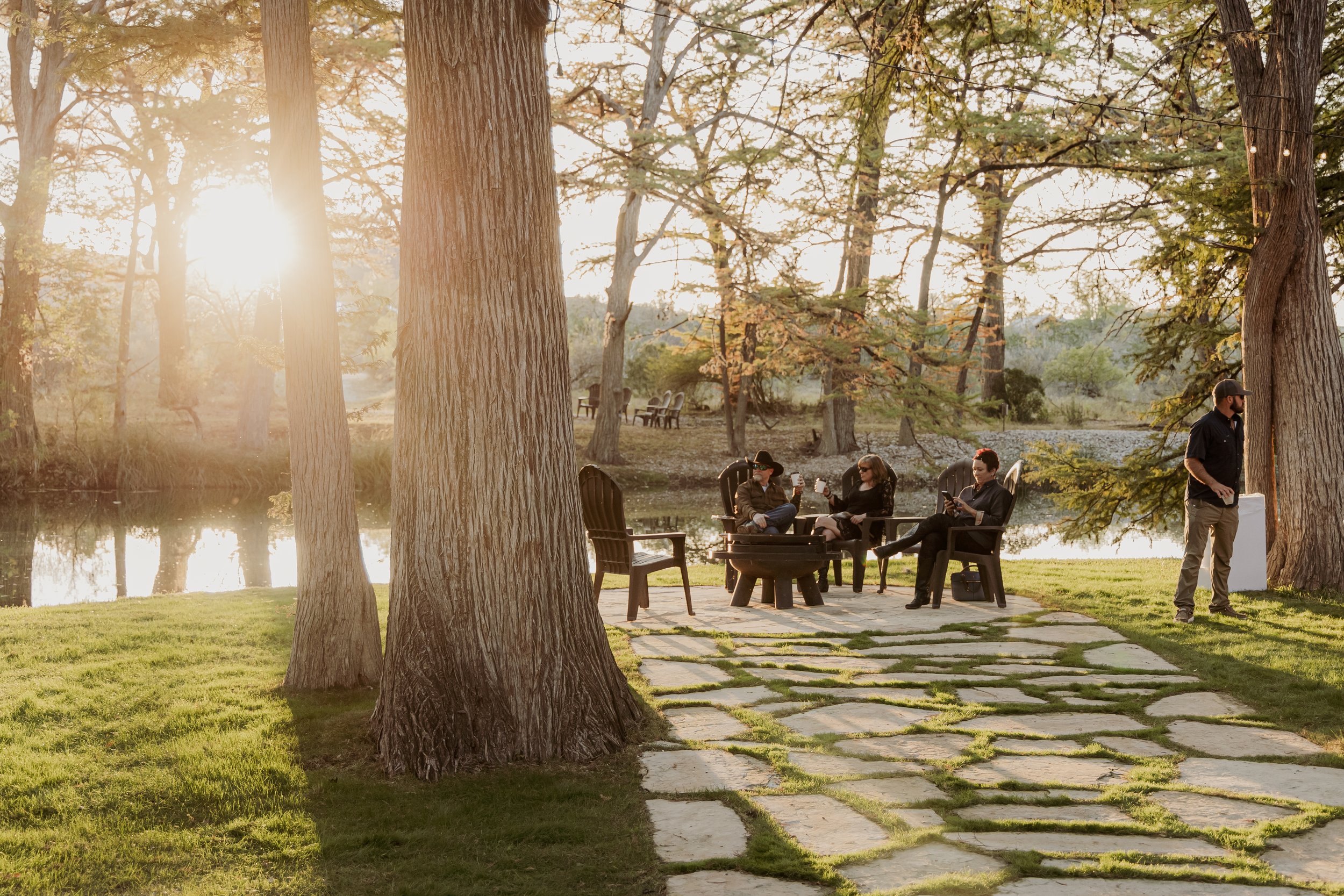 Four people sitting around a campfire in Adirondack chairs by a pond, with a man standing nearby, surrounded by trees during sunset.