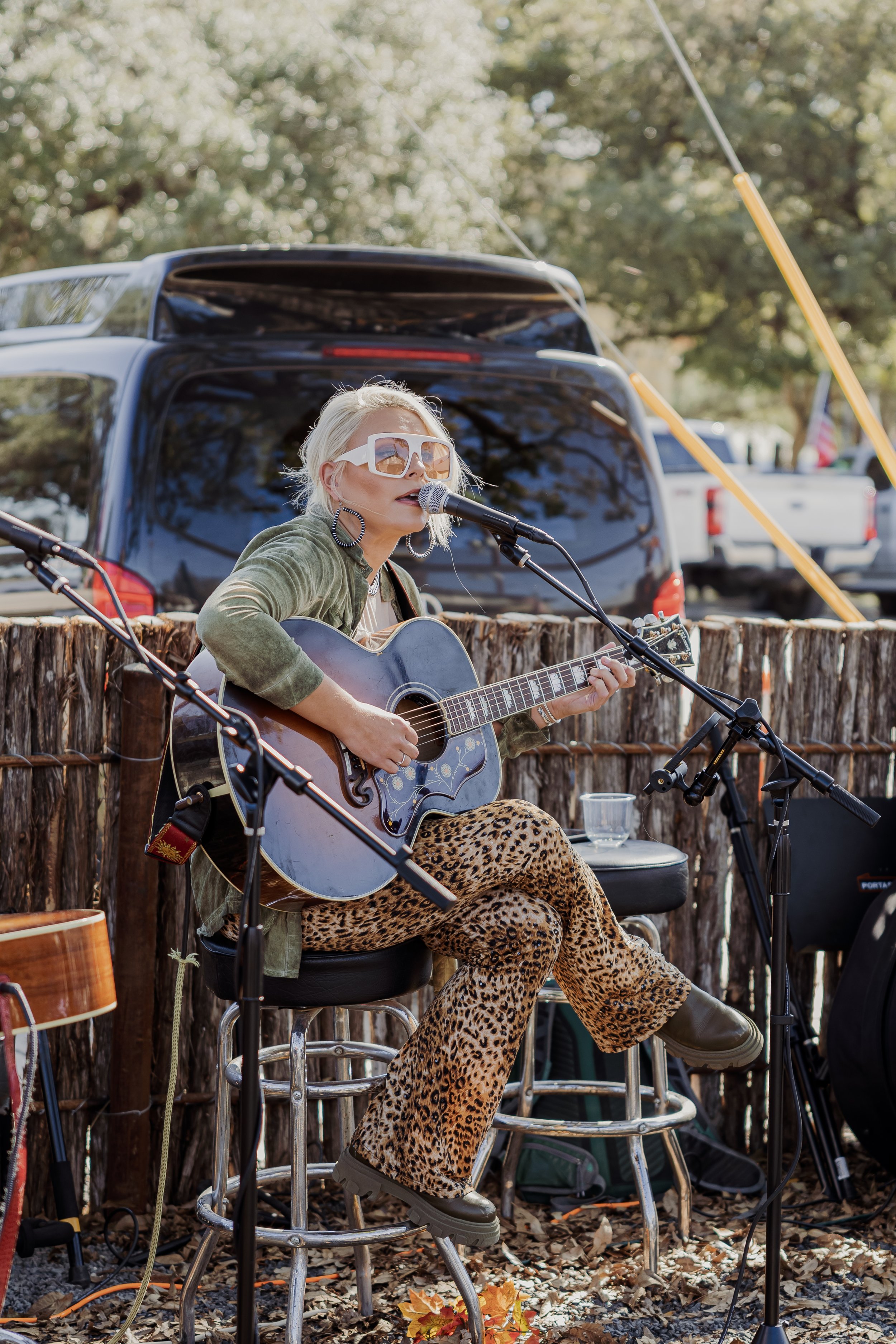 A woman with short platinum blonde hair, wearing large sunglasses, earrings, a green jacket, and leopard print pants, is playing an acoustic guitar and singing into a microphone outdoor at a rustic venue with trees and parked vehicles in the backgrou