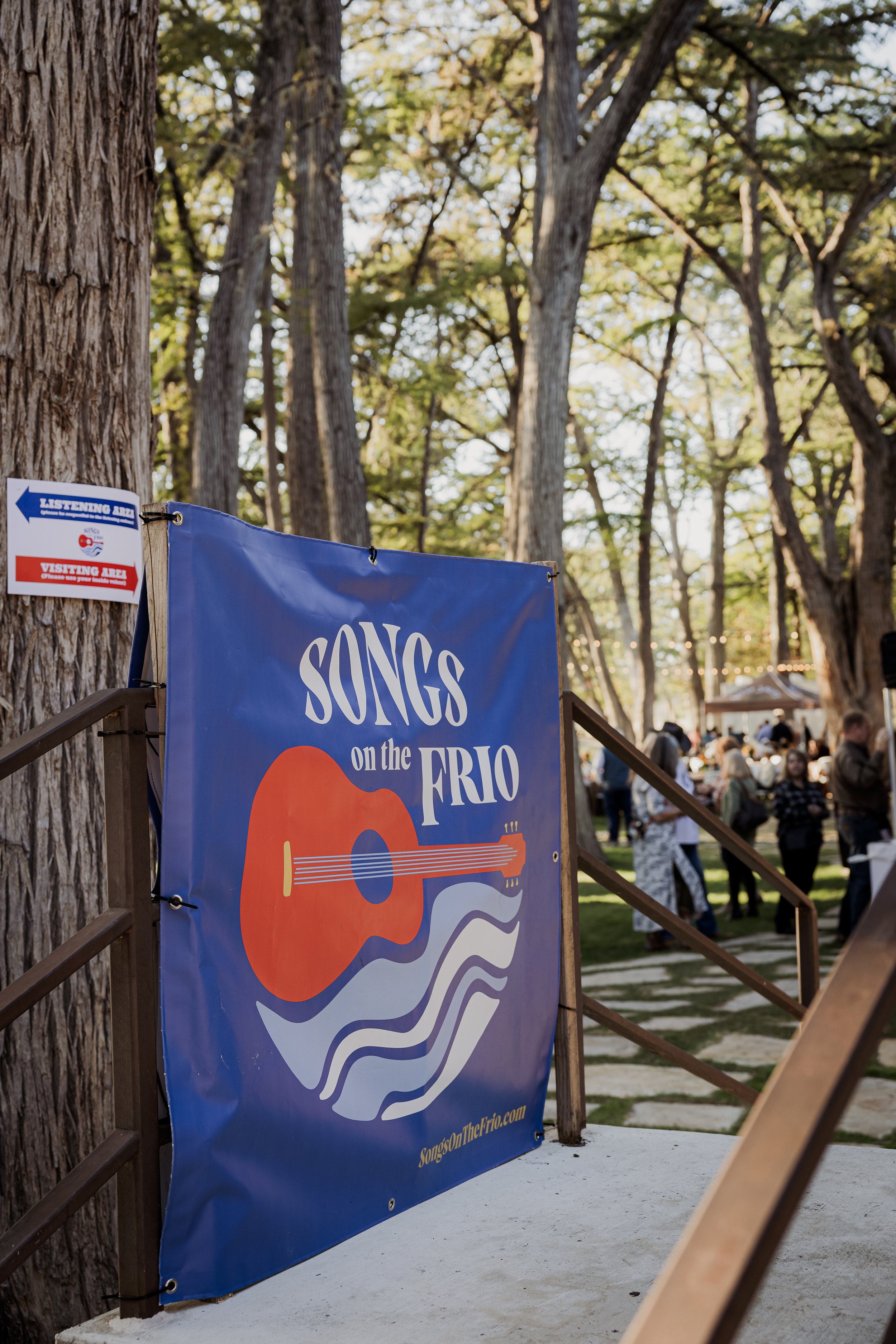 A blue festival banner reads "Songs on the Frio" with an illustration of a guitar and musical waves, set outdoors among trees, with people in the background at an outdoor event.