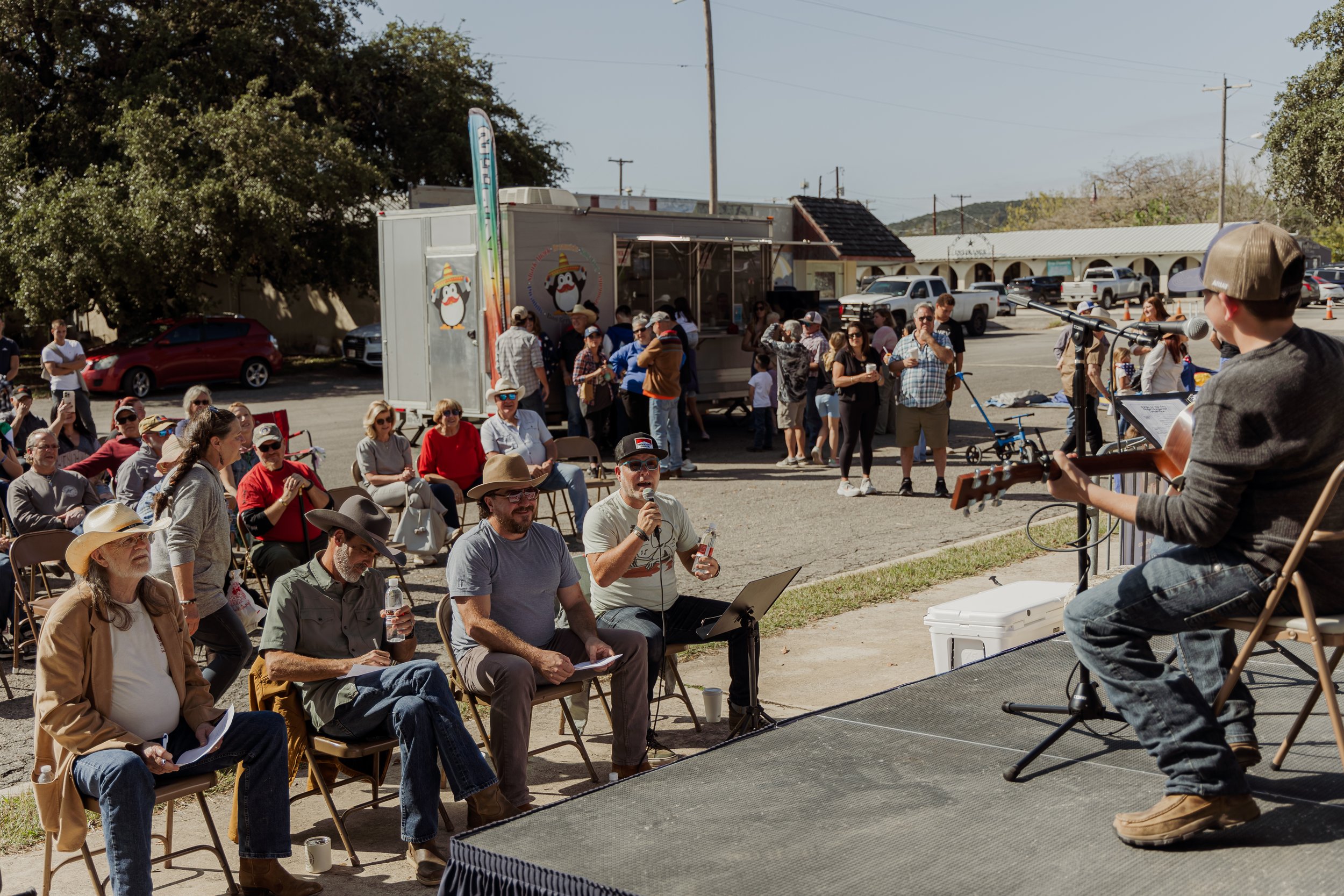 A young boy wearing a cap and gray sweater playing guitar and singing into a microphone at an outdoor event. An audience of mostly adults, some wearing cowboy hats and sunglasses, seated and standing, watching the performance. In the background, ther