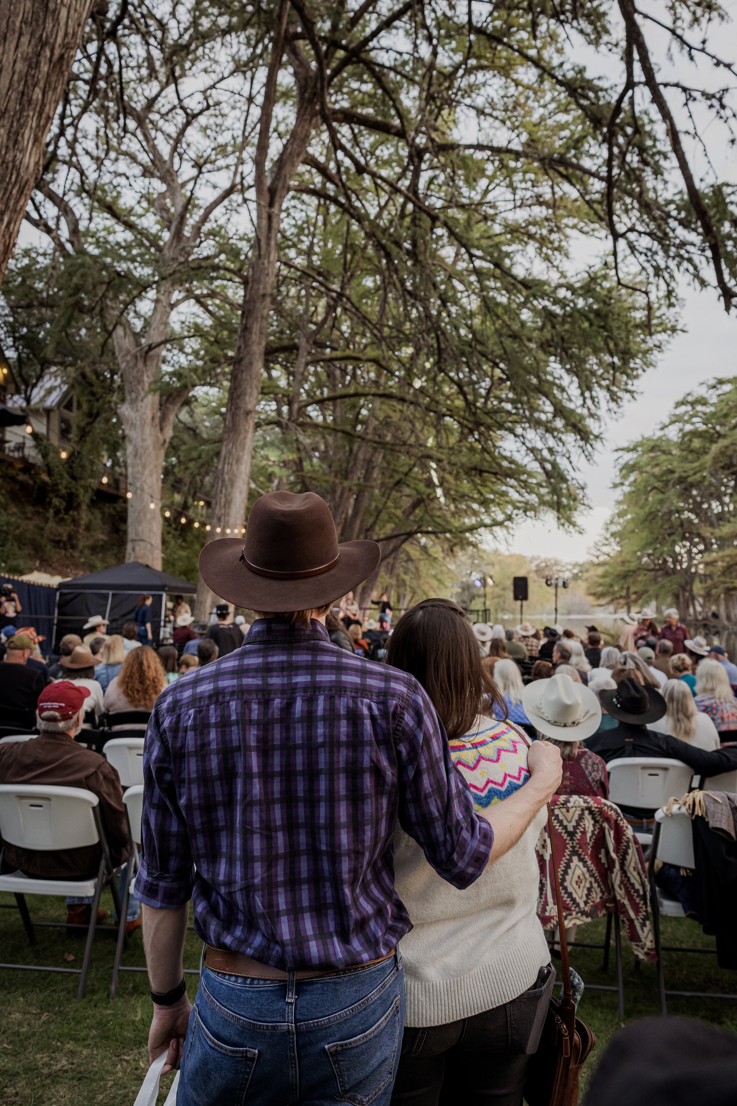 A crowd of people seated outdoors in a park-like setting, watching a stage performance among large trees with string lights.