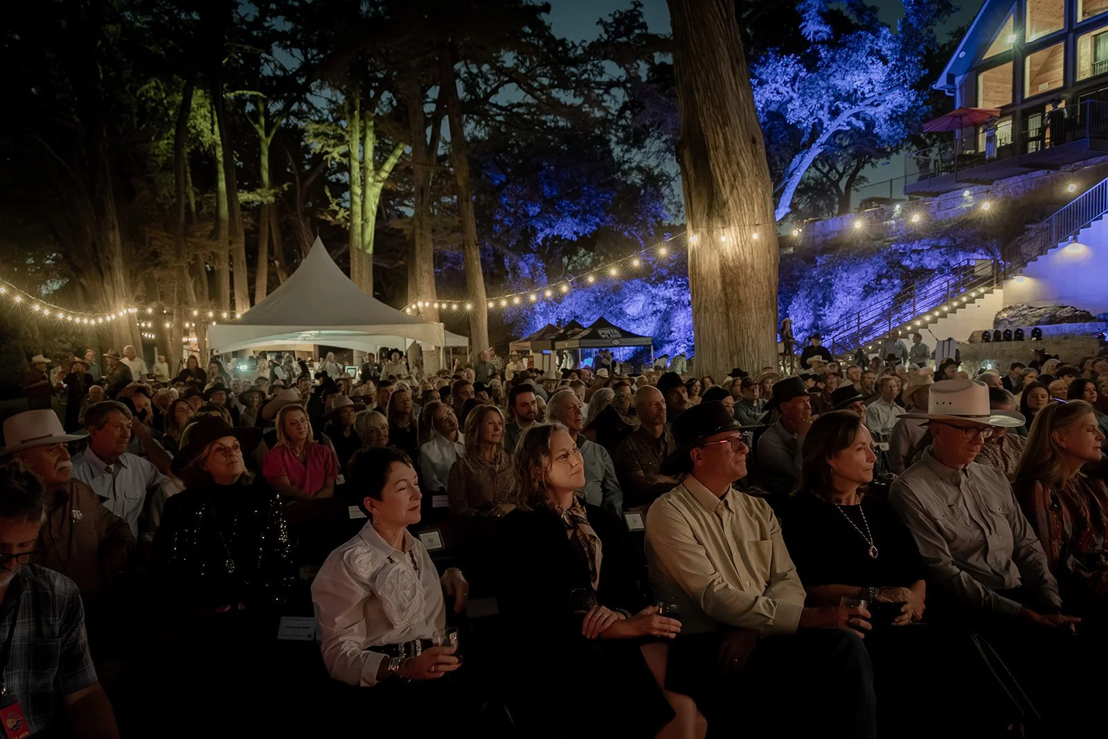 Crowd of people seated outdoors at night under string lights, surrounded by tall trees and illuminated purple-blue foliage, attending an event near a building with stairs and outdoor seating.