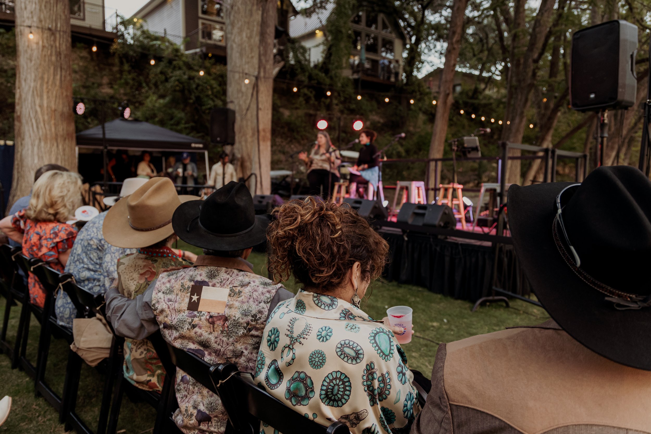 People watching an outdoor musical performance at dusk, seated on chairs in a wooded area with string lights overhead.