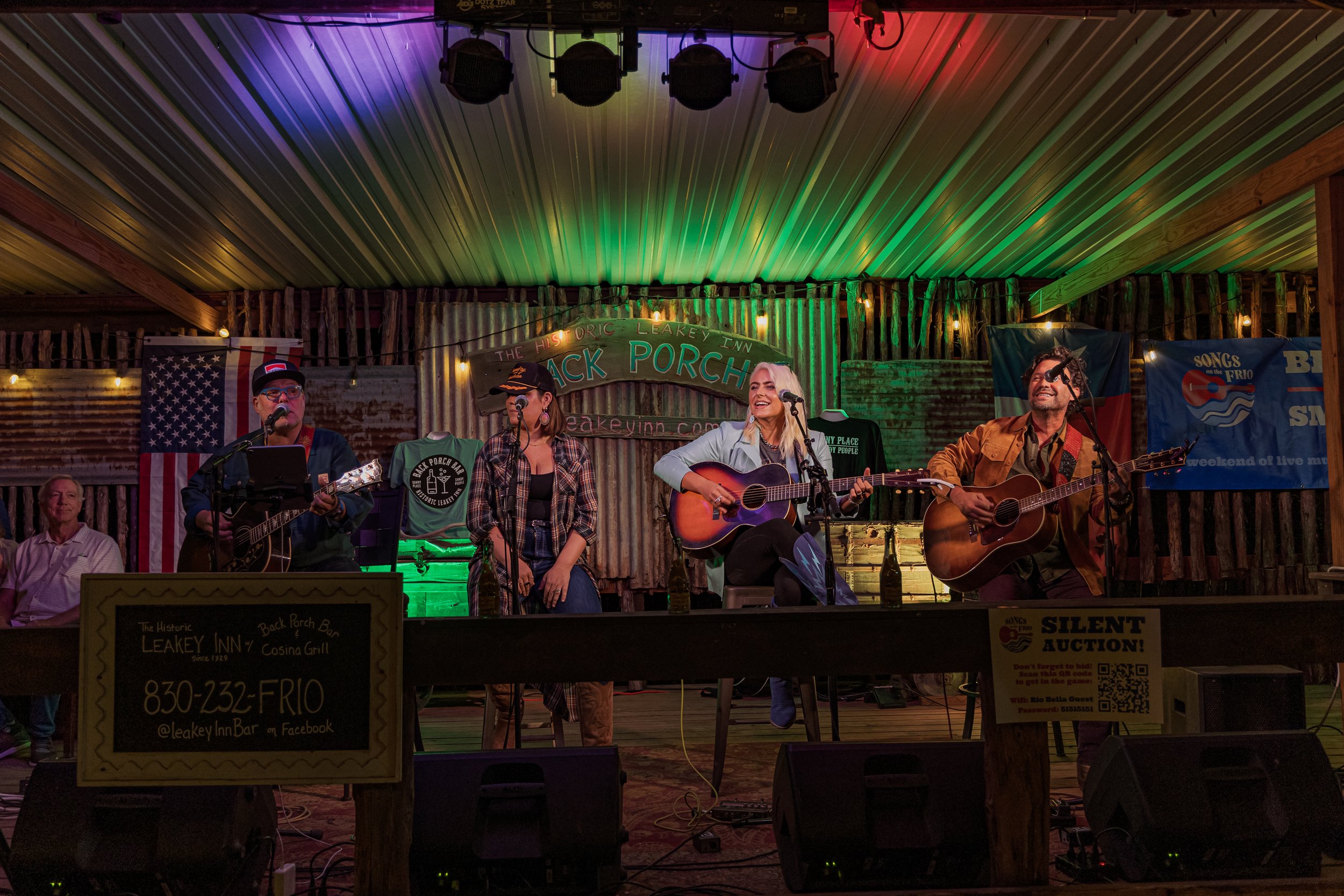 A band of four musicians performing on stage at a rustic venue, with country-style wooden decorations and banners in the background, and colorful stage lighting.