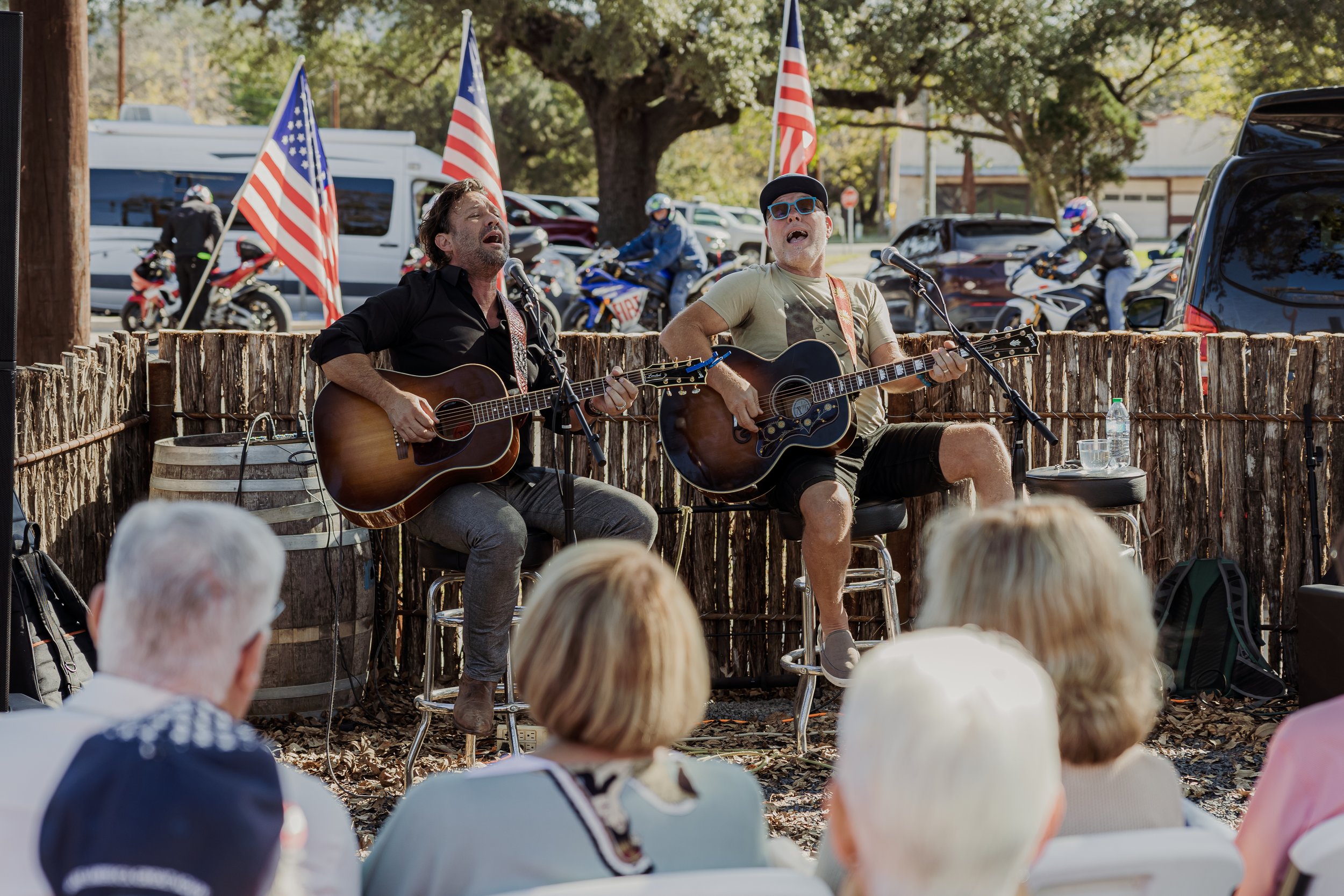 Two musicians playing acoustic guitars and singing on an outdoor stage with American flags in the background, audience watching.