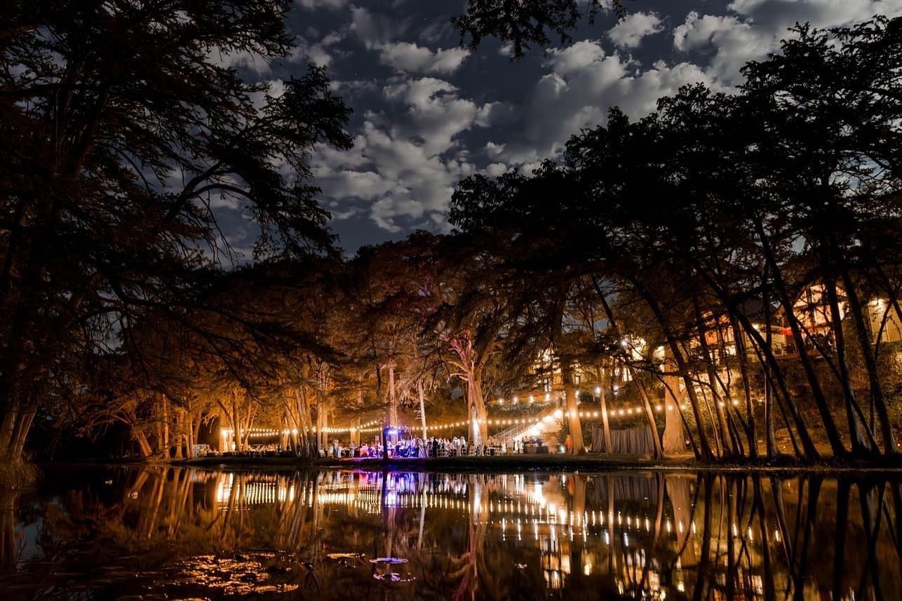Nighttime outdoor music festival event by the Frio River with string lights, trees, and a cloudy sky, reflected in the water at Rio Bella Resort.