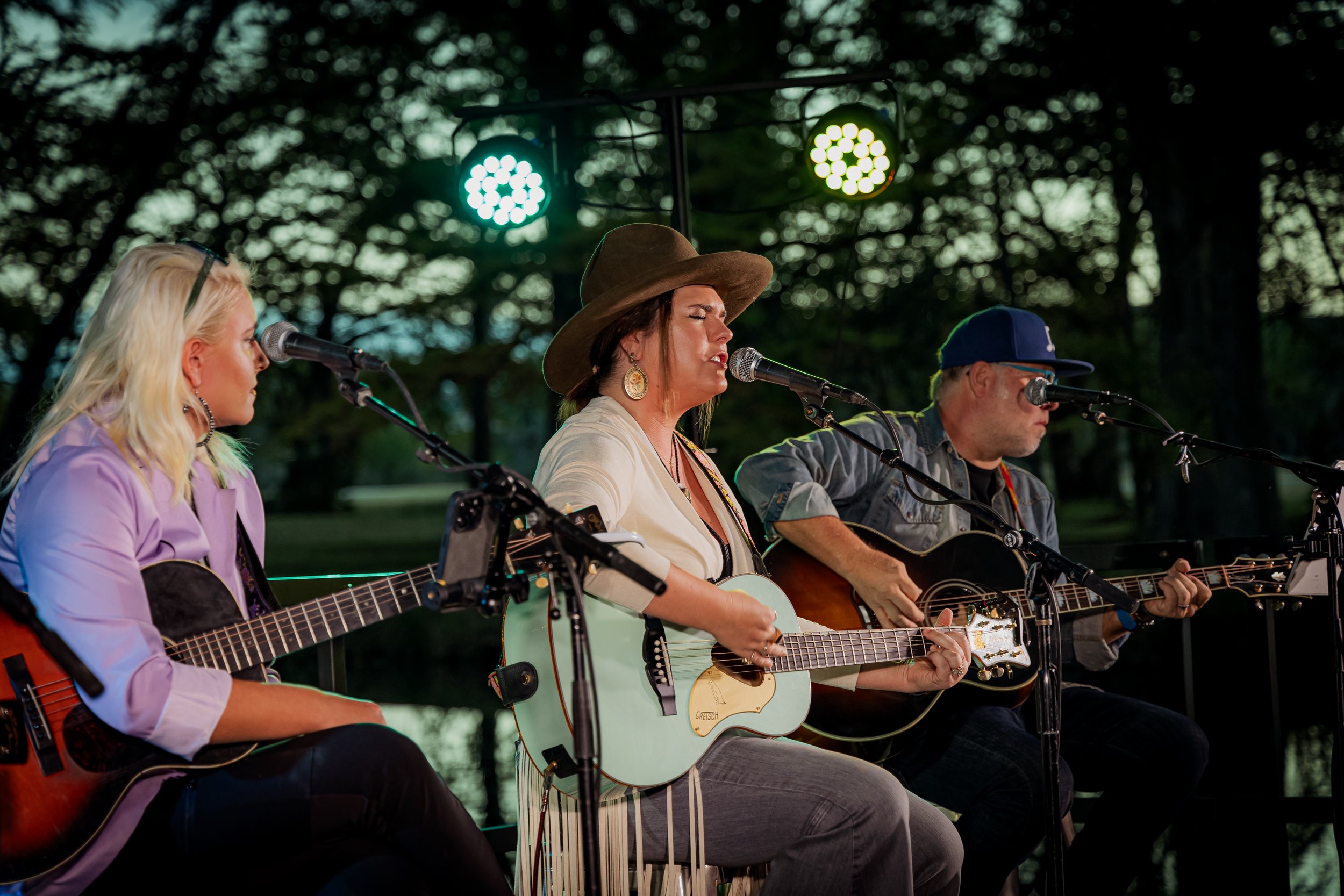 Three musicians performing outdoors at sunset, playing acoustic guitars and singing into microphones, surrounded by trees with green lights overhead.