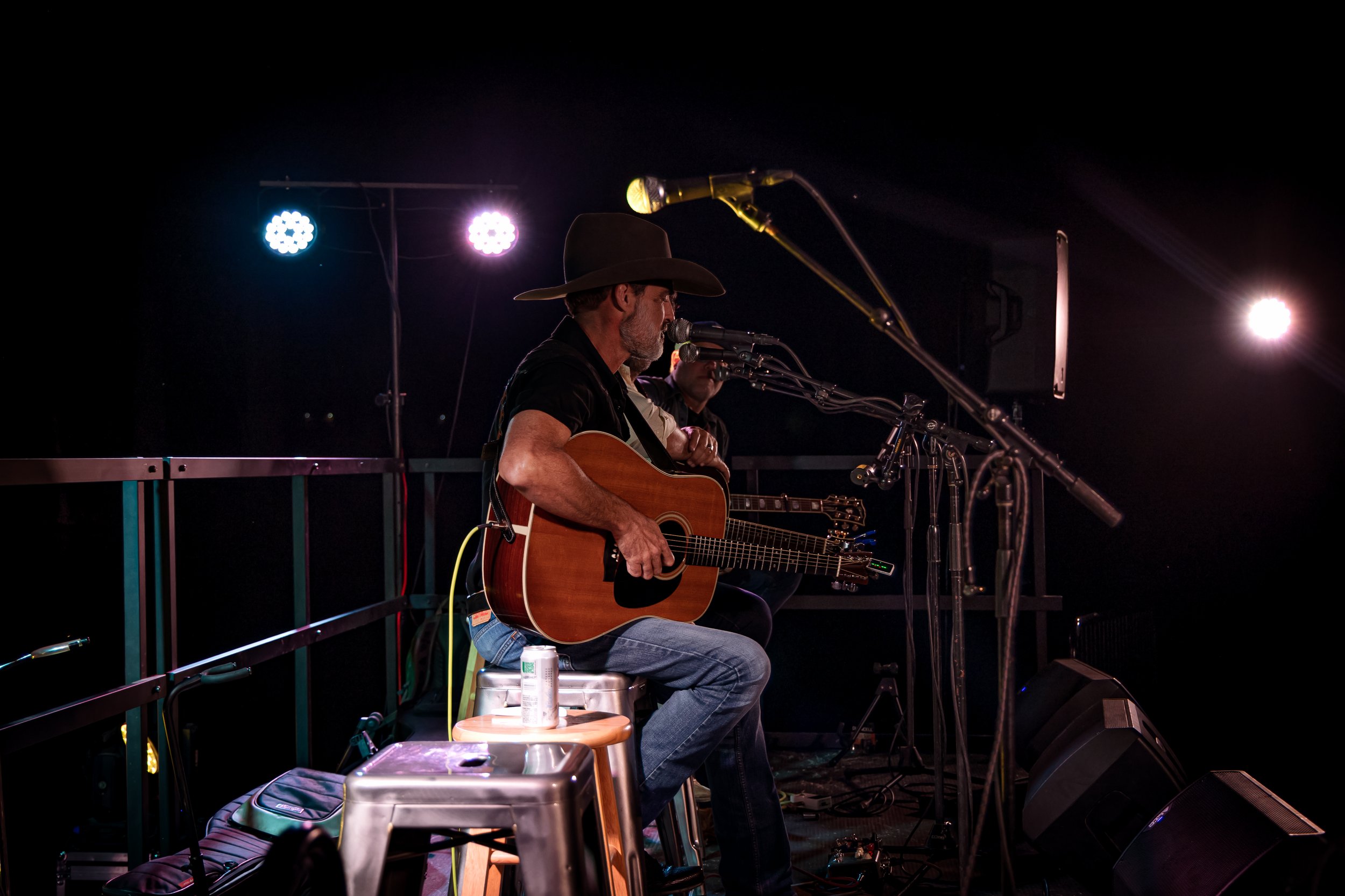 Musician in cowboy hat playing acoustic guitar on stage with microphones, stage lights, and sound equipment.