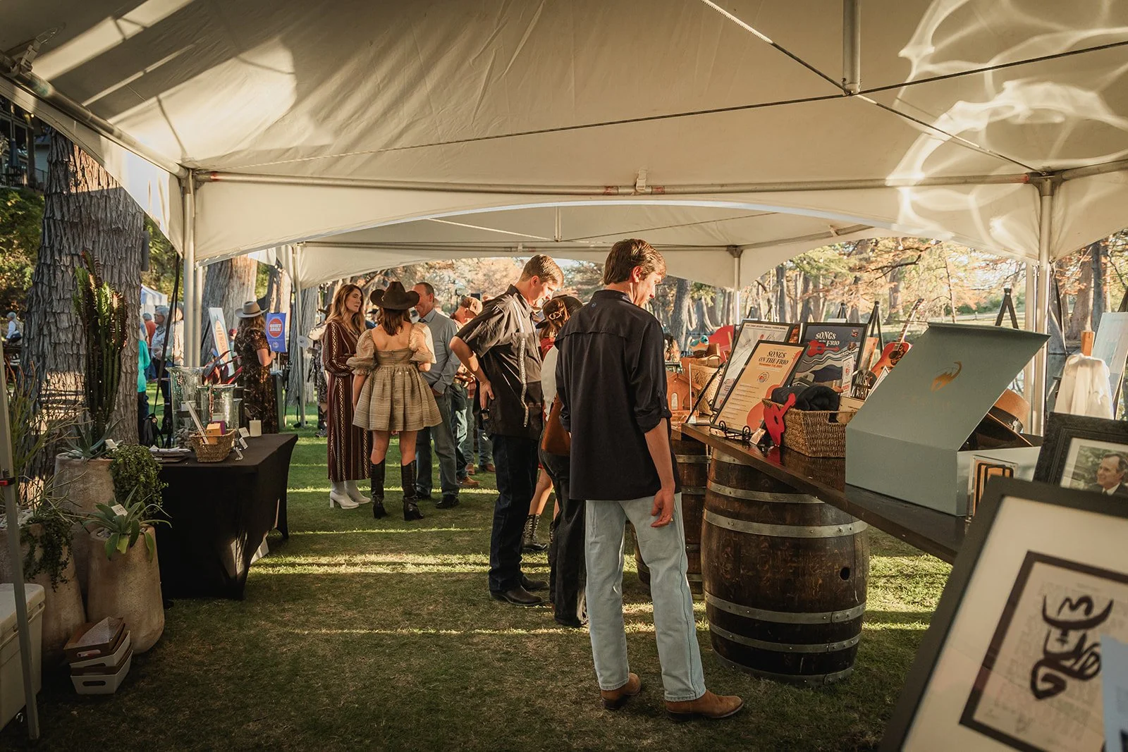 People shopping at an outdoor marketplace or fair, browsing various items displayed on tables under a white canopy, with trees and other vendors visible in the background.
