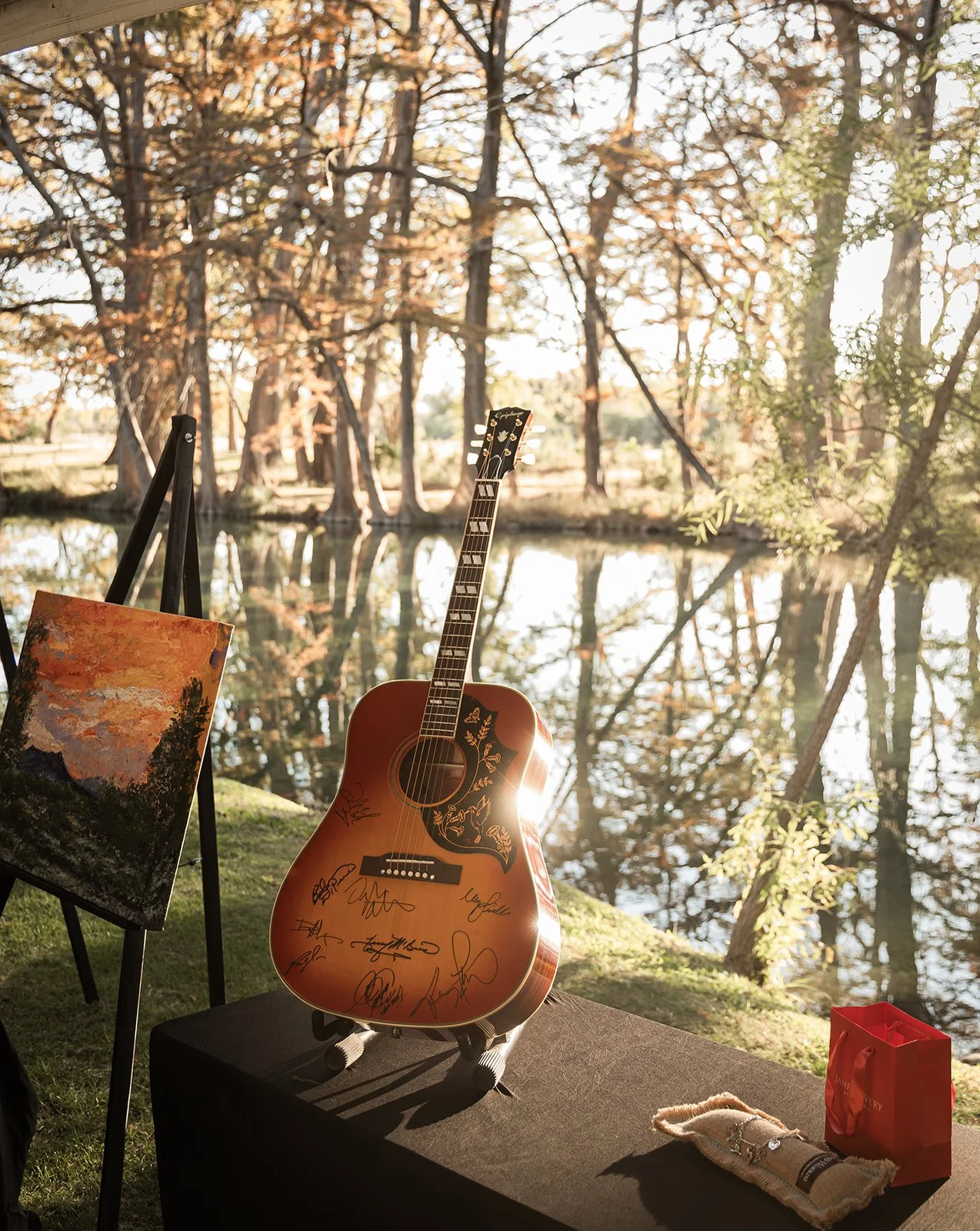 An autographed guitar resting on a table outdoors near a lake, with a painting on a stand and a red gift bag nearby, during sunset or sunrise with trees reflected in the water.