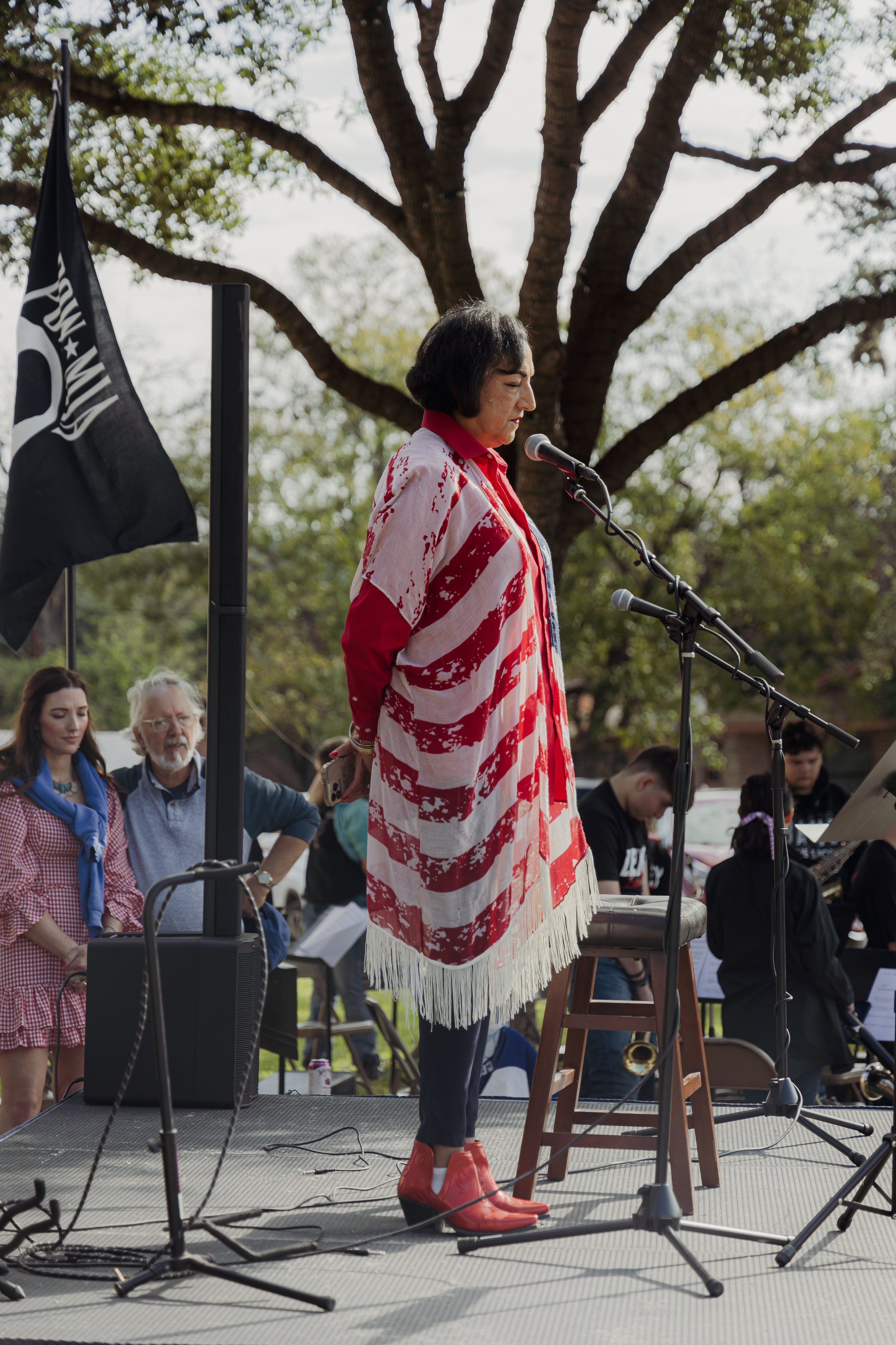 A woman stands on a stage with a microphone, wearing a red, white, and blue poncho and red boots, with people gathered behind her at an outdoor event.