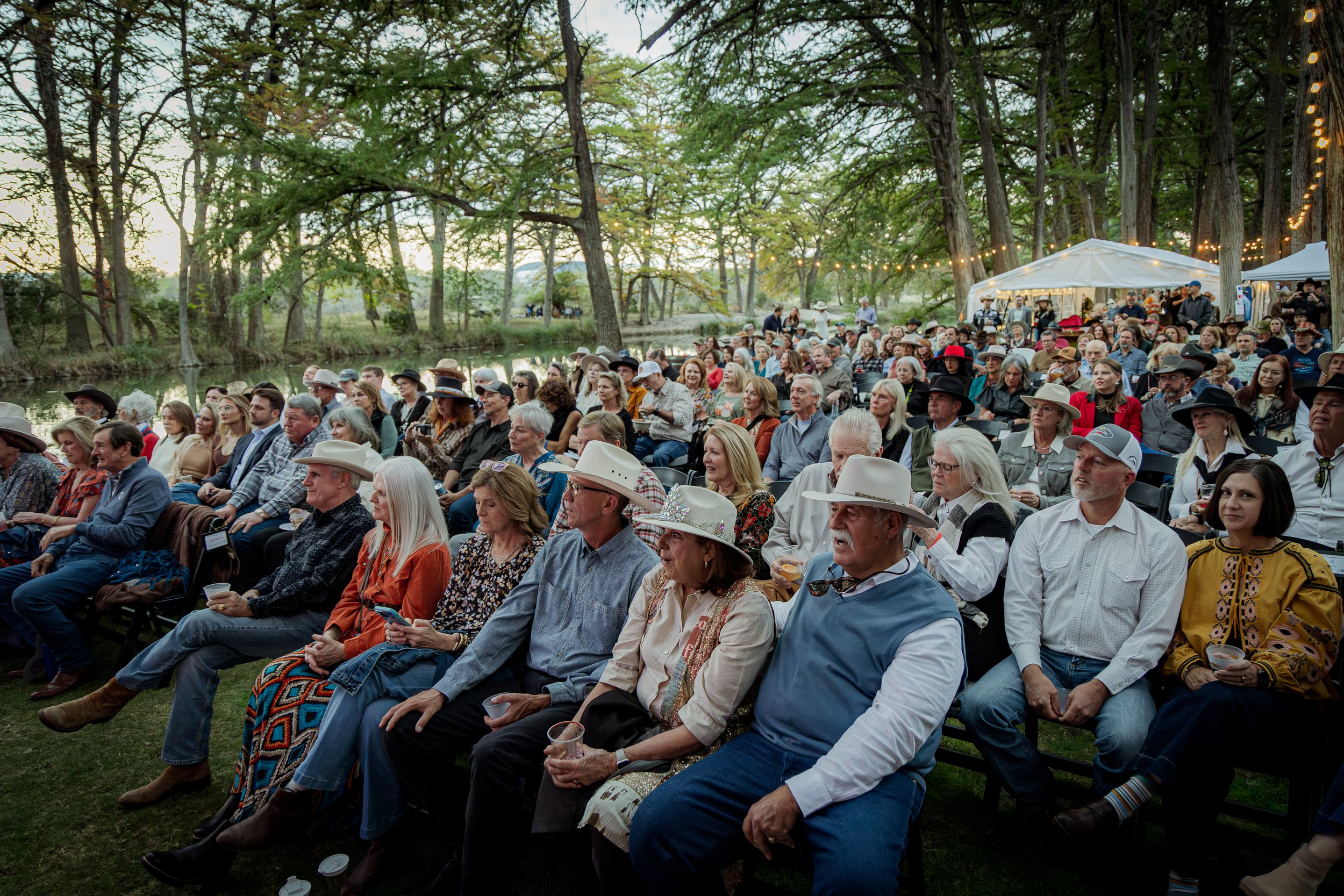 A large outdoor gathering of people seated near a small body of water, attending an event during dusk, with string lights and trees surrounding the area.