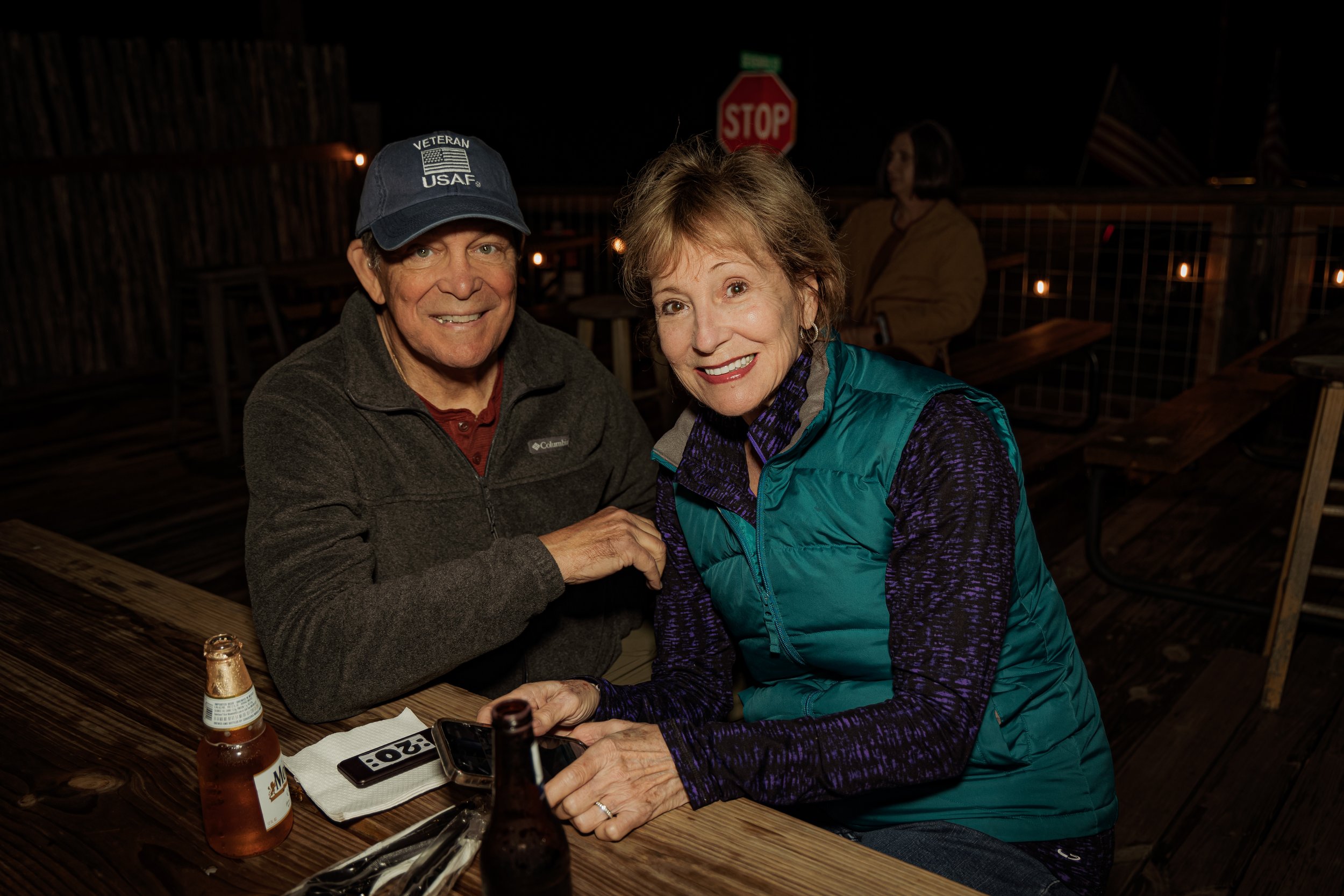 A smiling man and woman sitting at a wooden table outdoors at night, with drinks and a phone in front of them.