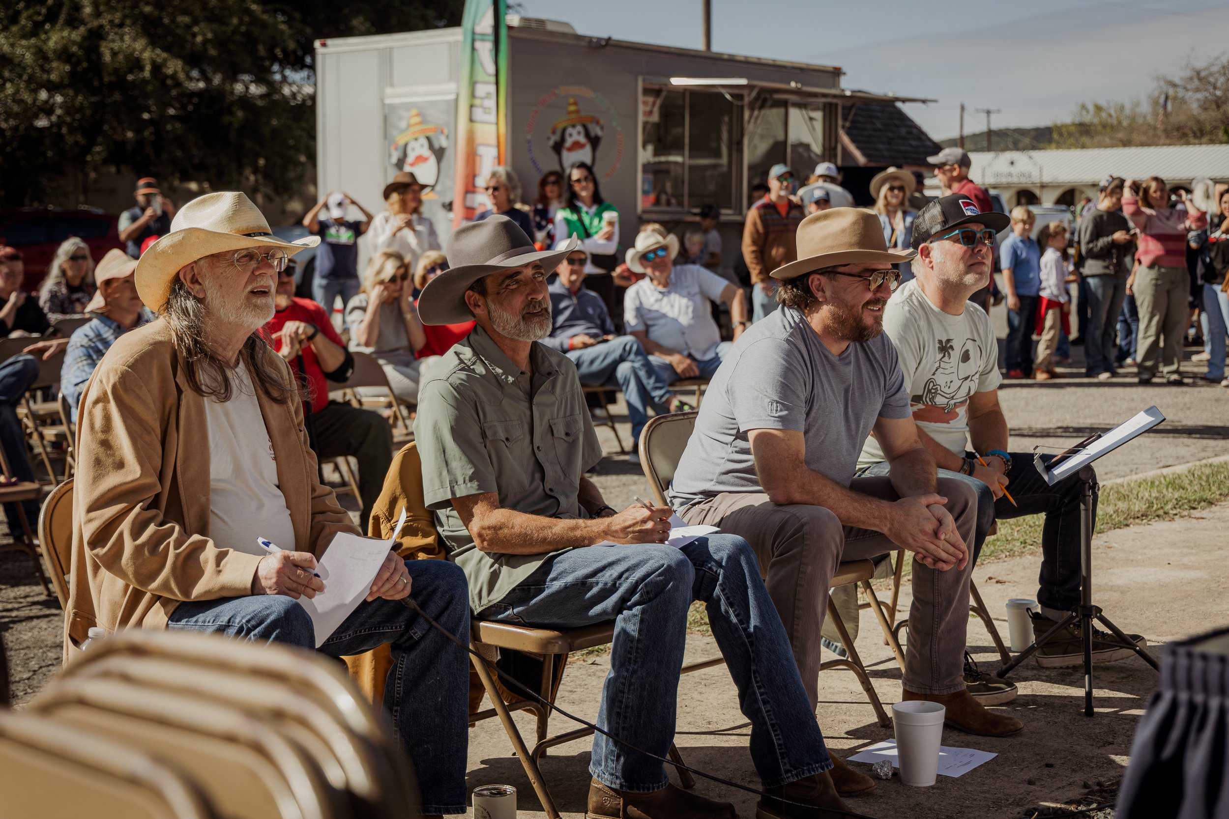 A group of four men seated outdoors at a public event, with a crowd of people standing and sitting behind them, some wearing hats and sunglasses, and a food truck visible in the background.