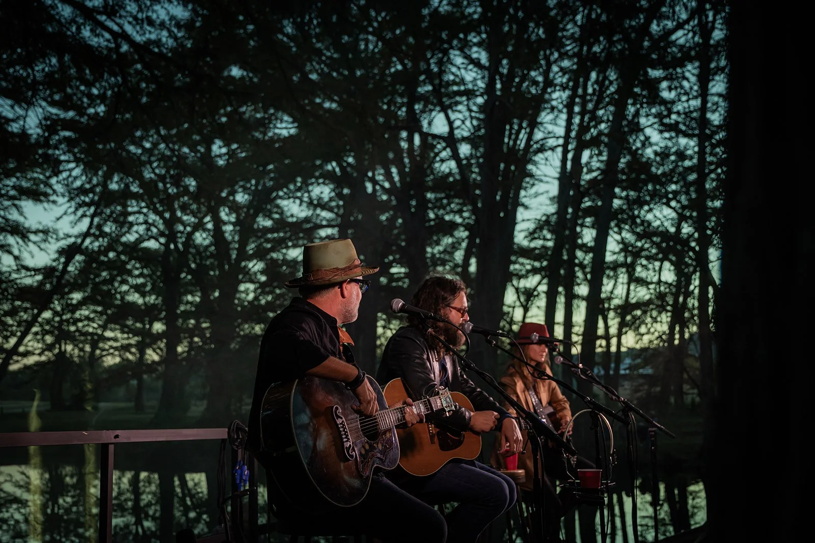 Three musicians performing outdoors at sunset, playing guitars and singing into microphones, seated in front of a wooded background.
