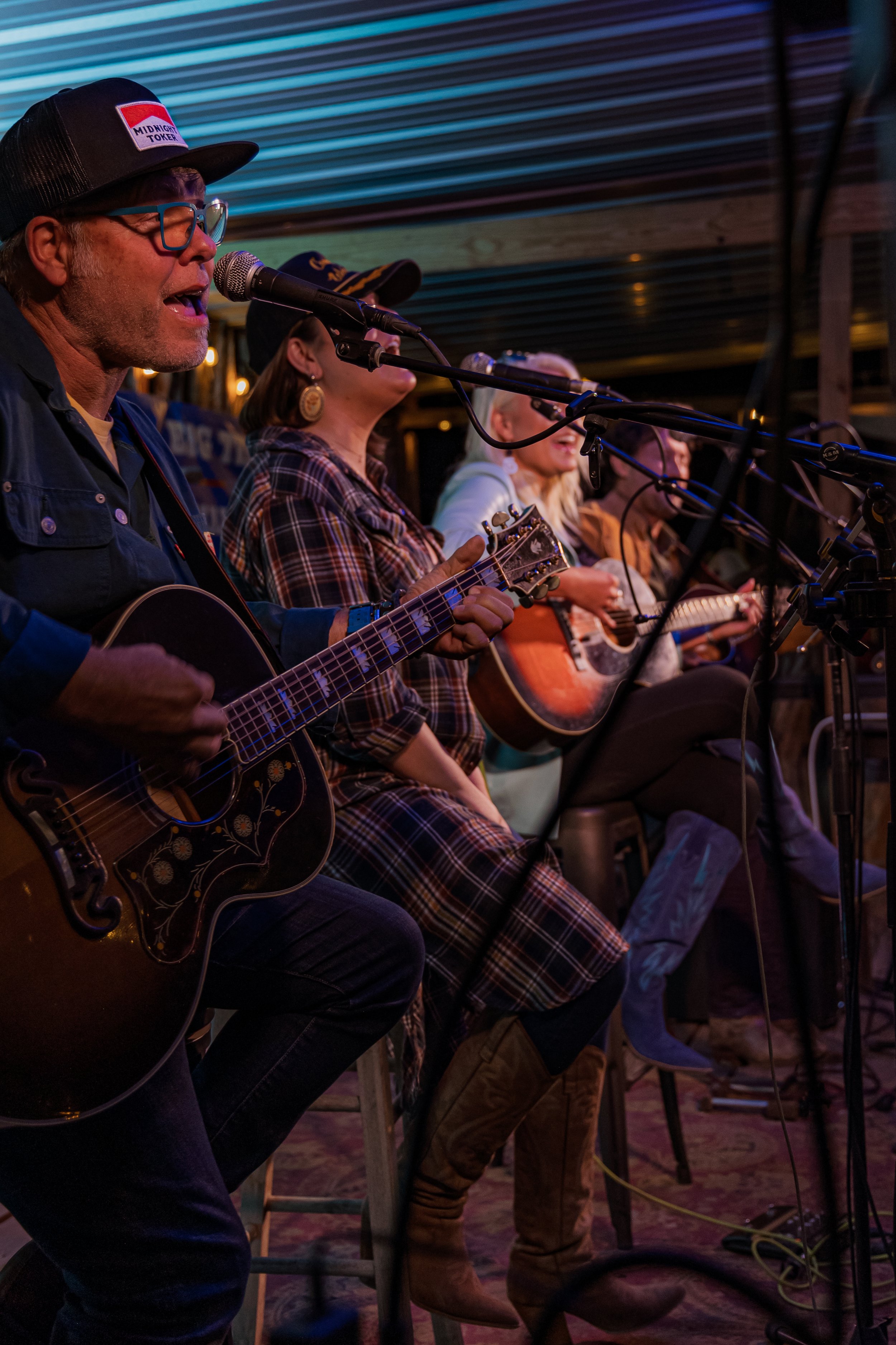 A group of people sitting on stools, playing acoustic guitars and singing into microphones at an indoor music event.