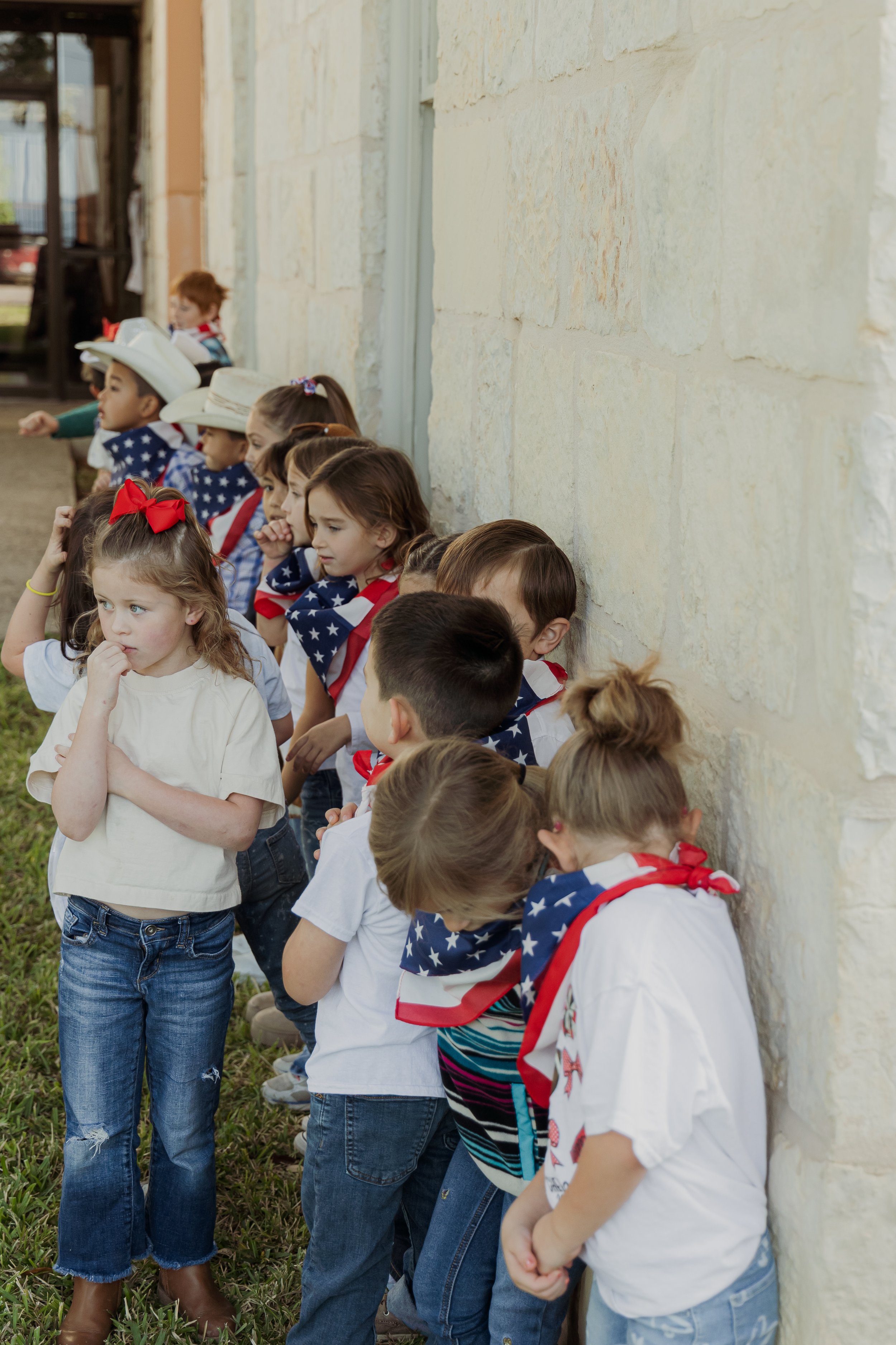 Children standing against a stone wall, some wearing American flag bandanas, suggesting a patriotic event or celebration.