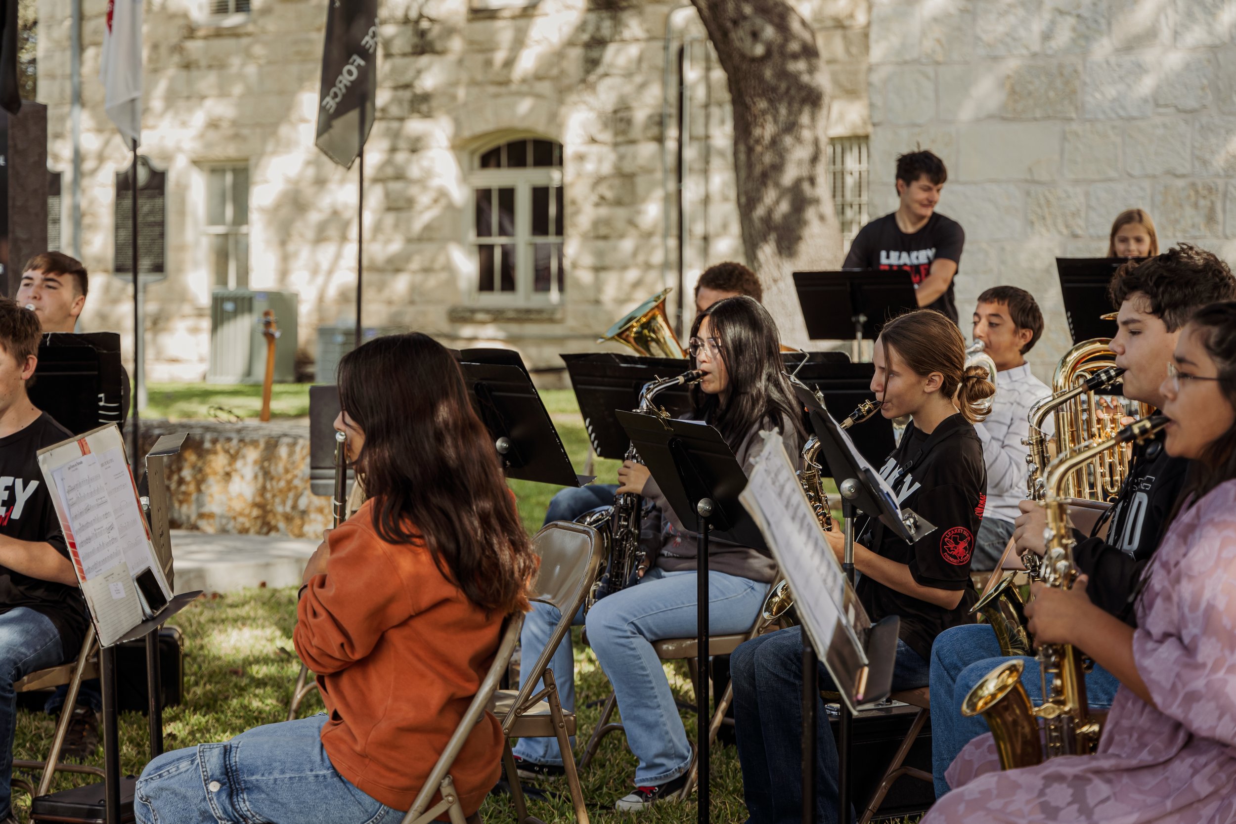 A group of young musicians playing wind instruments outdoors, seated on chairs with music stands, with a stone building and a large tree in the background.