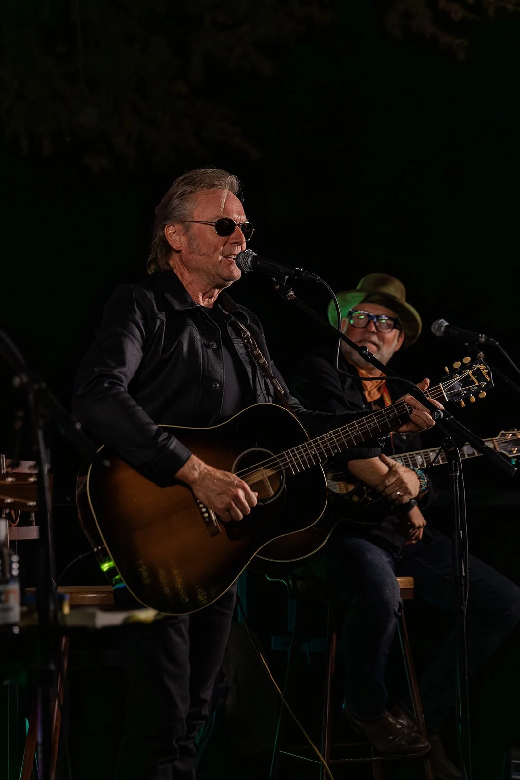 Two musicians performing on stage, one singing and playing acoustic guitar, the other sitting nearby with an acoustic guitar, both wearing dark clothing and hats, at an indoor music event.