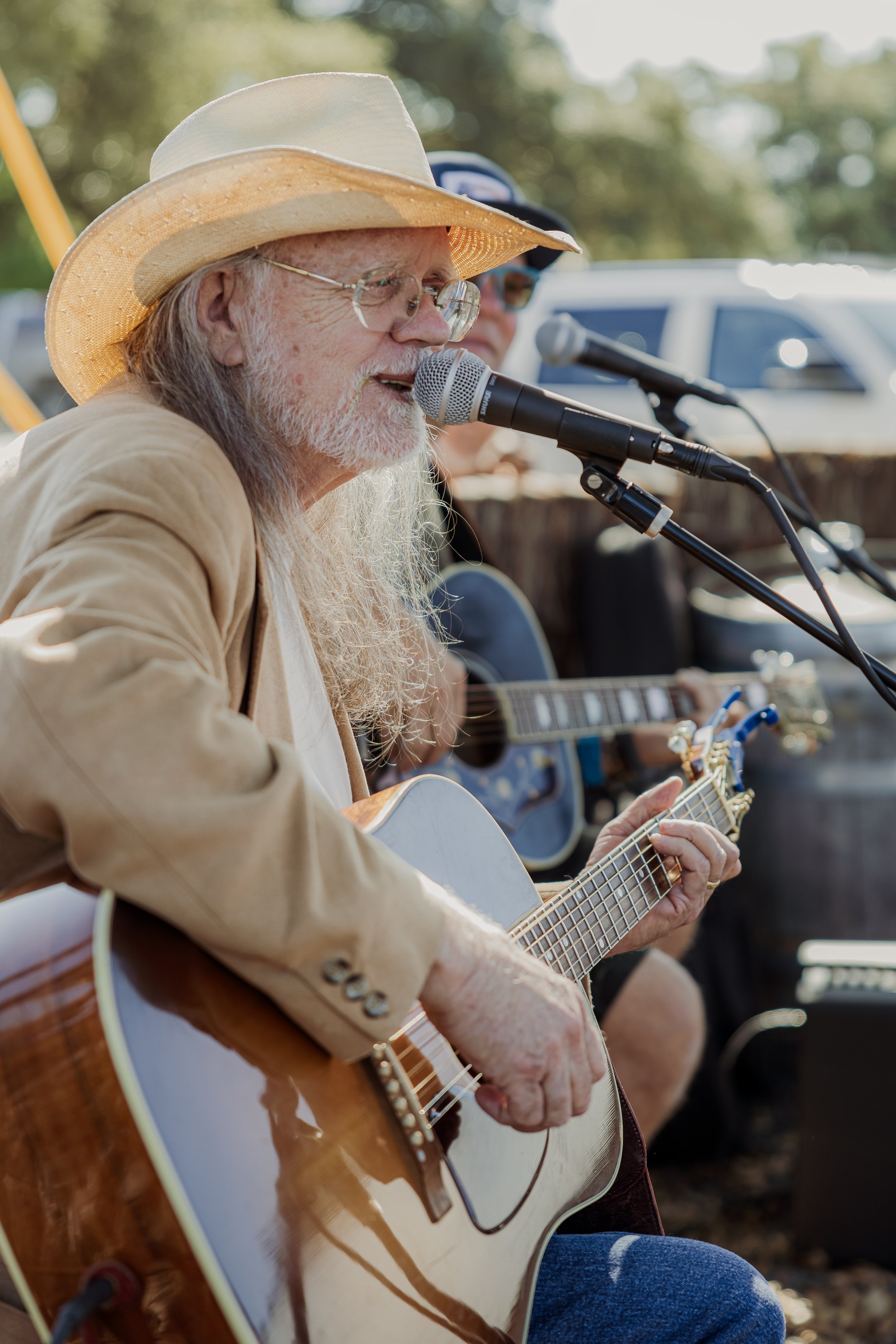 An older man with long gray hair, a beard, glasses, and a straw hat, playing an acoustic guitar and singing into a microphone at an outdoor event.