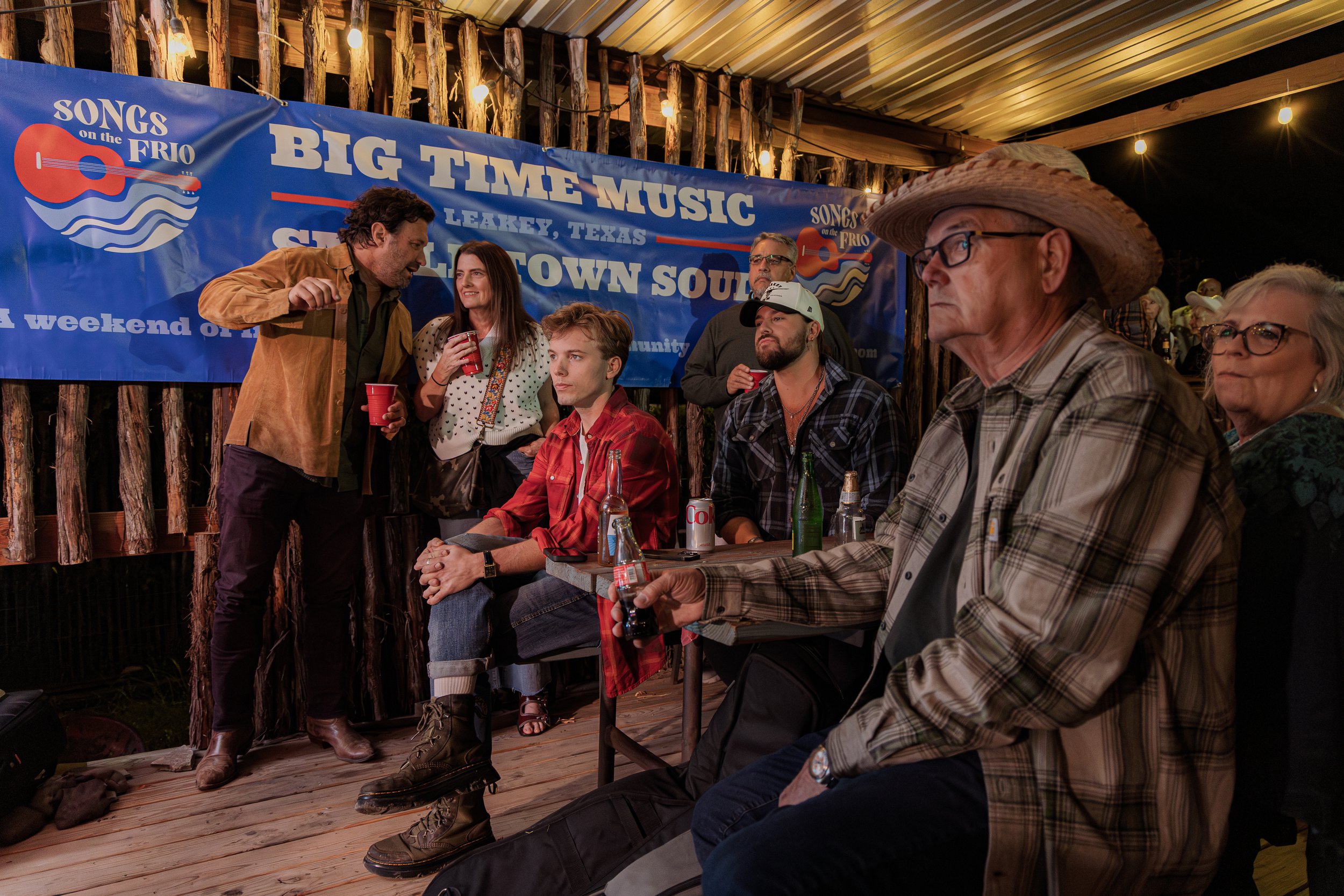 A group of people at a concert or festival, sitting at tables and standing outdoors under string lights, with a blue banner in the background that reads 'Big Time Music' and 'Songs on the Frio', indicating a country or folk music event in Leakey, Tex