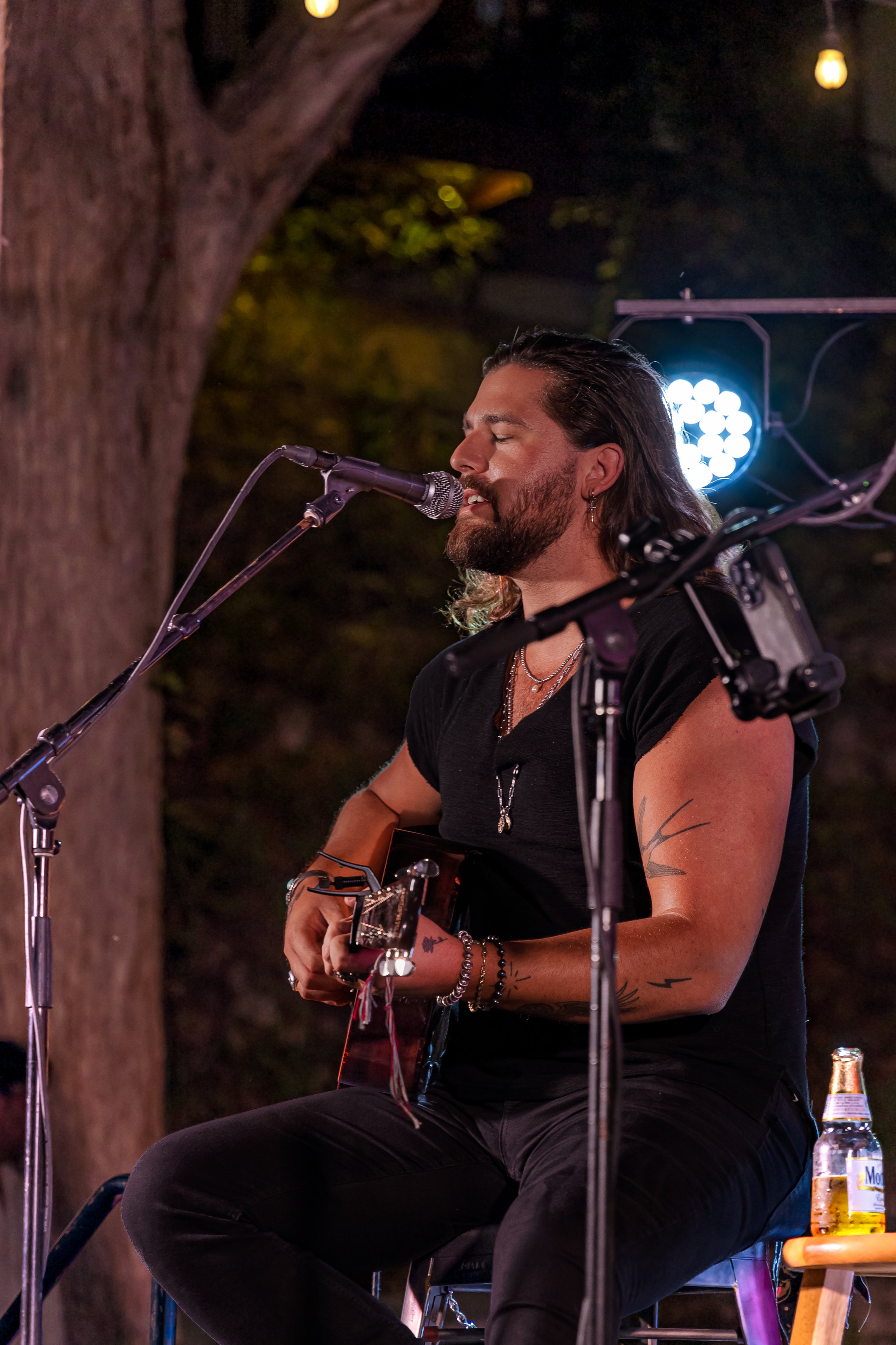A man with long hair and a beard playing an acoustic guitar and singing into a microphone during a nighttime outdoor performance, illuminated by stage lights.