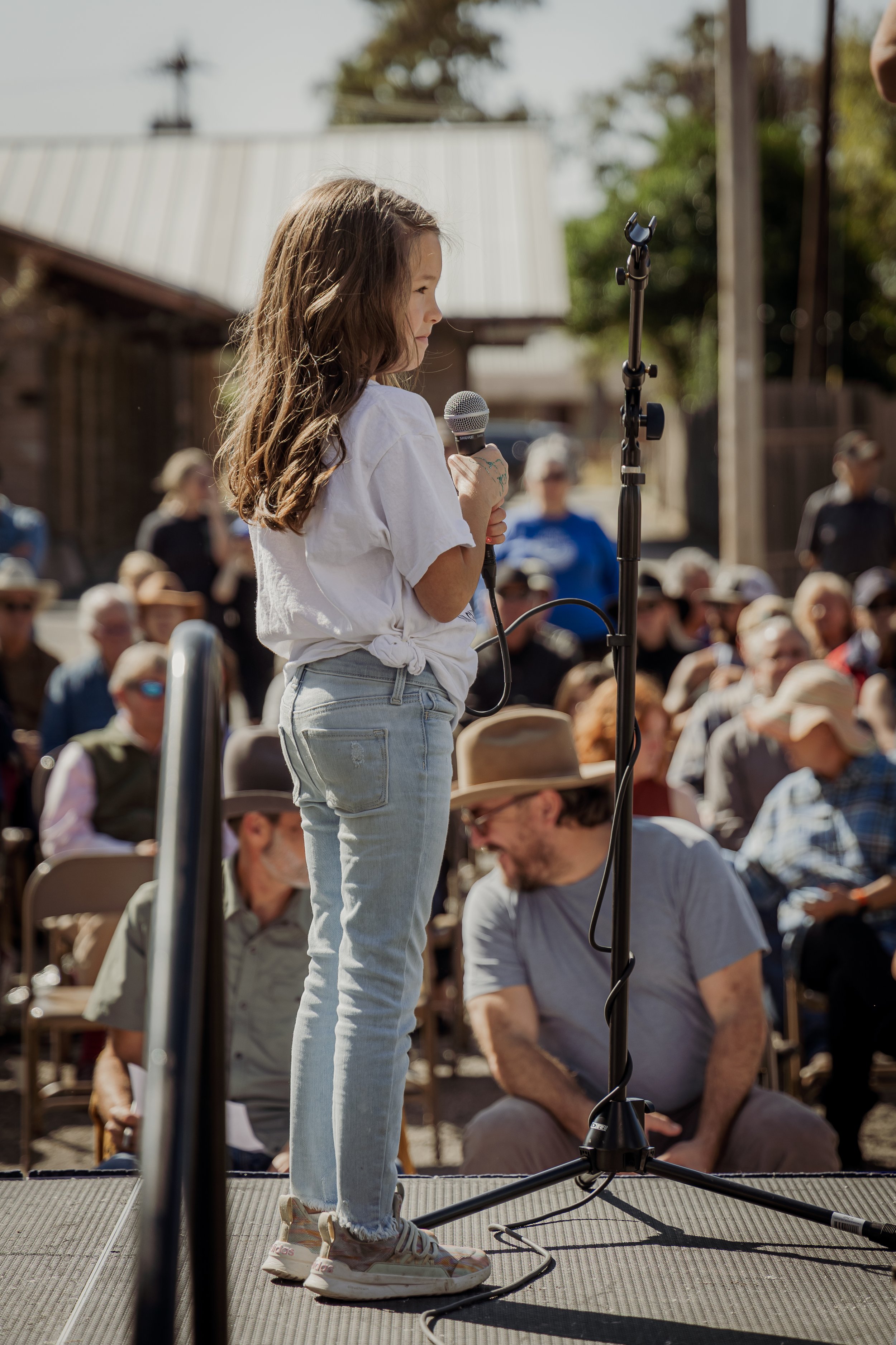 Young girl speaking into a microphone on stage at an outdoor event with a seated audience and a metal railing visible.