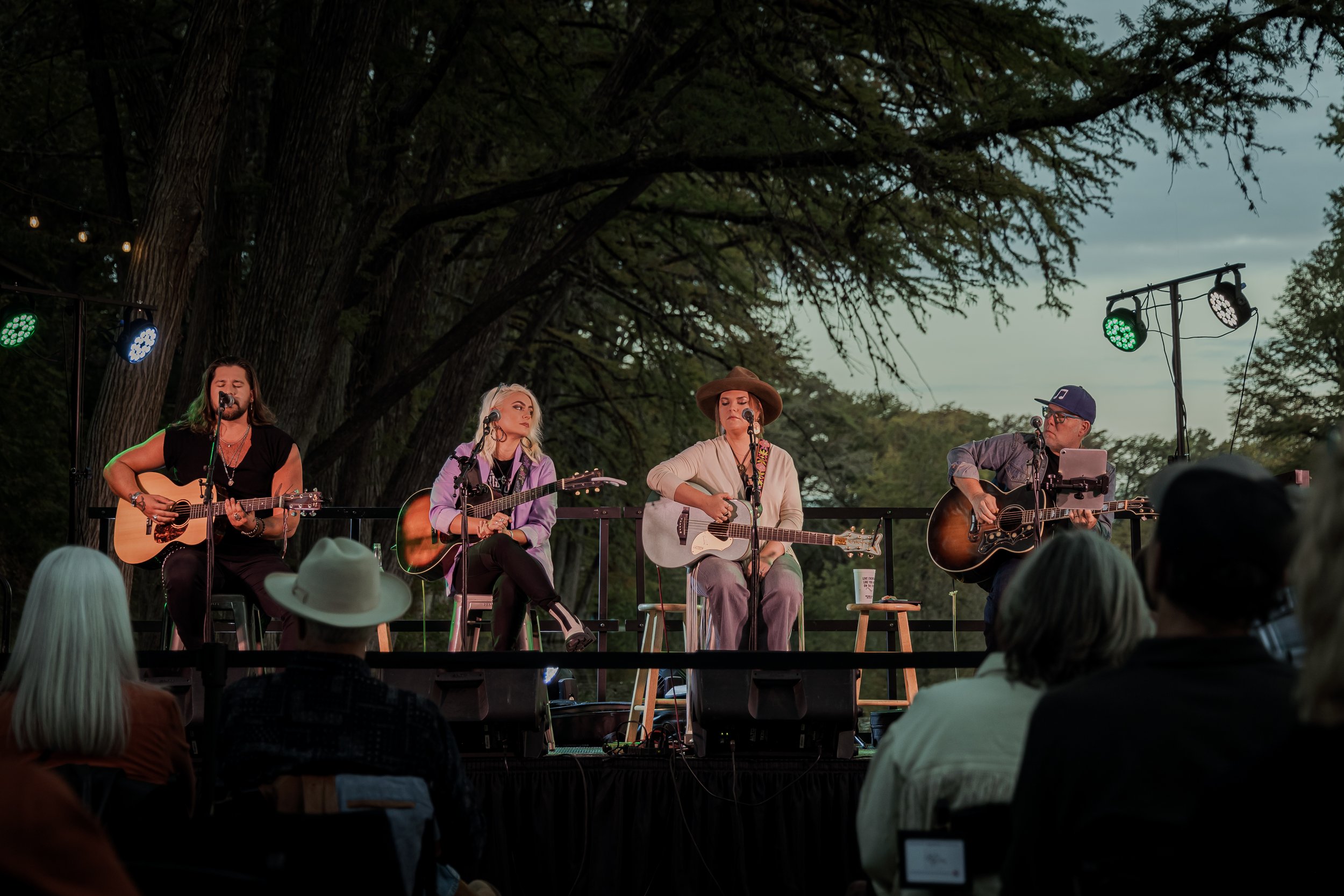 Four musicians performing on an outdoor stage under large trees, each playing a guitar and singing into microphones. The audience is seated in front, some with hats. The stage has colored lights and microphones, with a cloudy sky in the background.