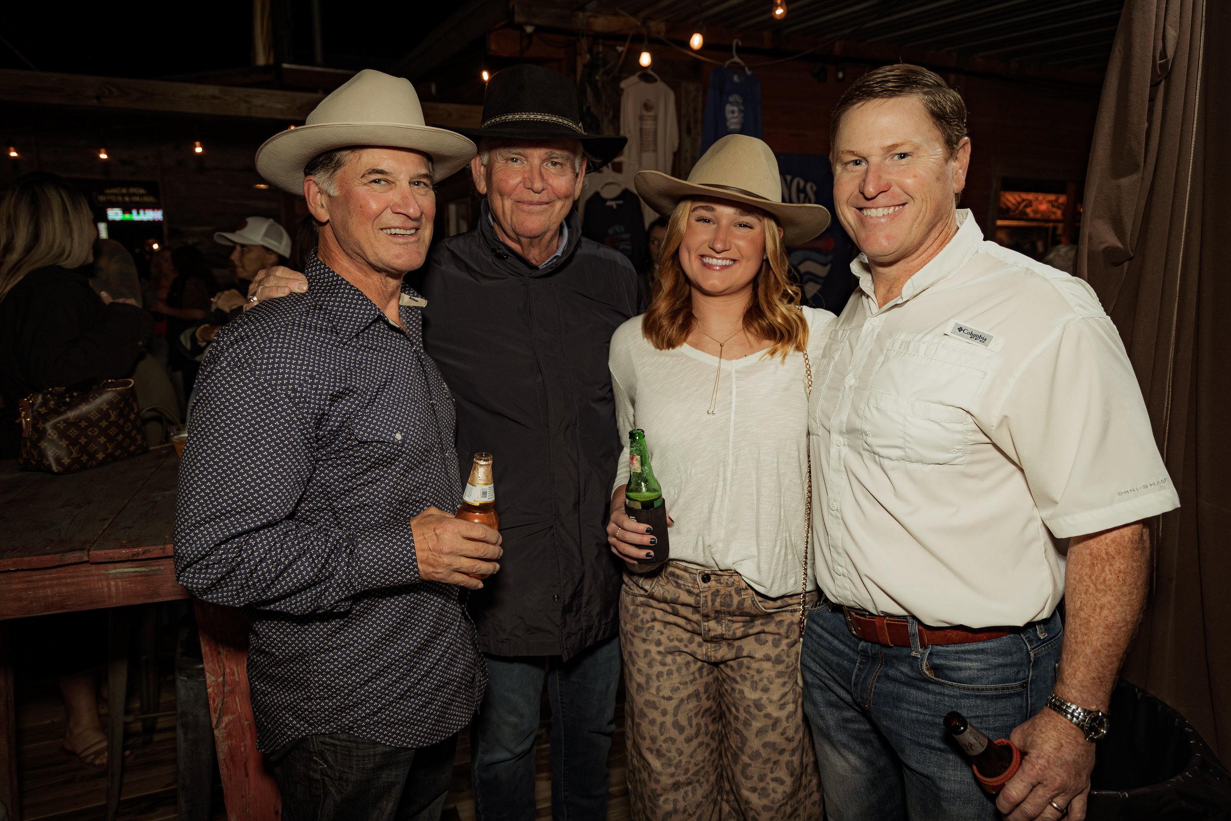 Group of four friends at a bar wearing cowboy hats, laughing, and holding drinks.